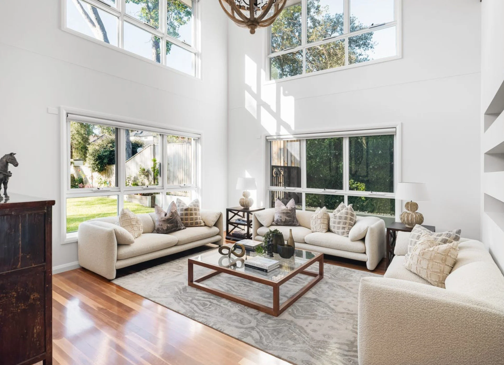 Bright living room with high ceilings, large windows, and white walls. There are three white sofas with patterned pillows, a wooden coffee table with decorative vases and books, a patterned area rug, and side tables with lamps. Outside, a green yard 