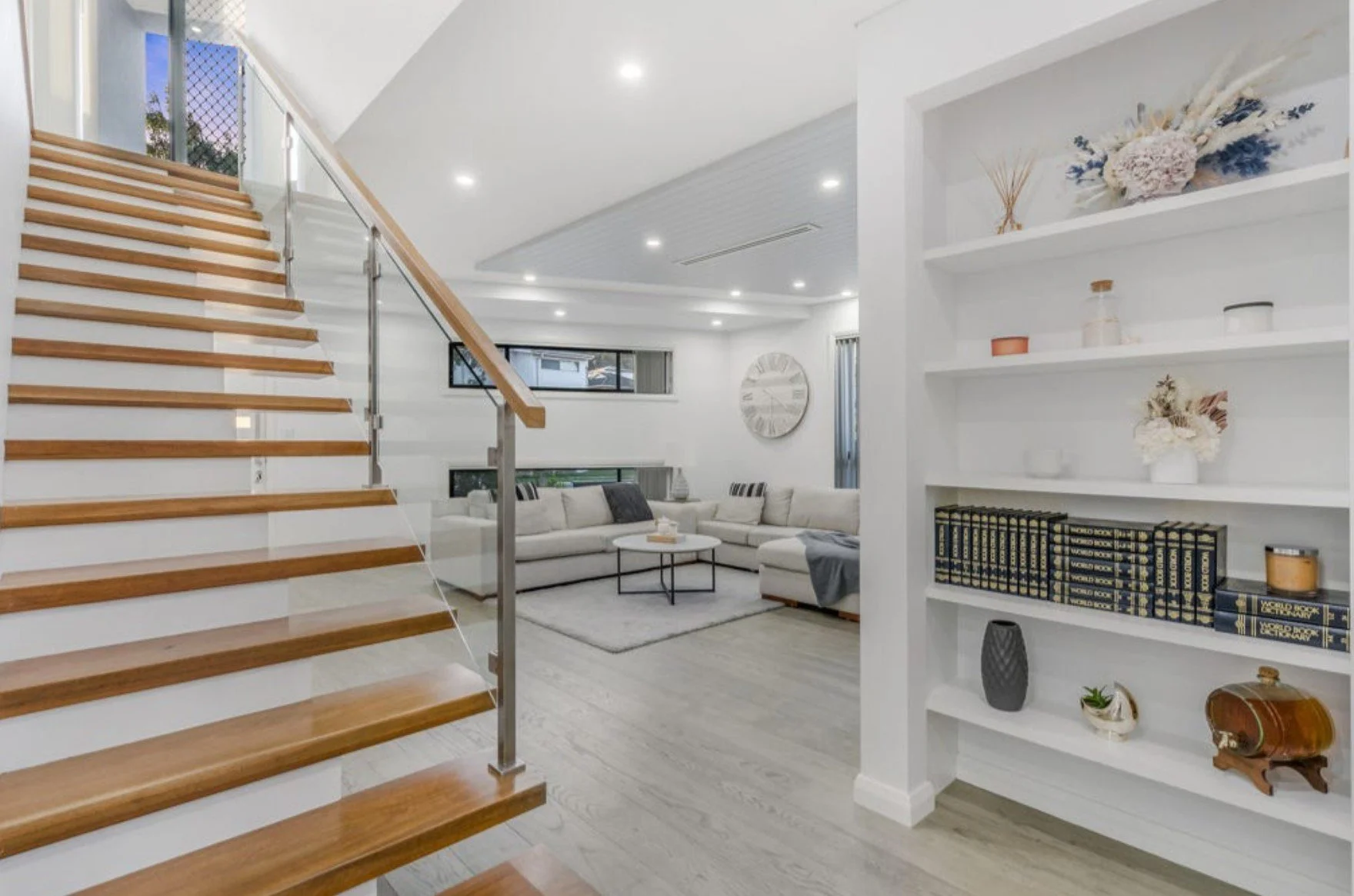 Modern living room with white walls, sectional sofa, coffee table, wooden stairs with glass railing, built-in bookshelf with decorative items, and large windows.