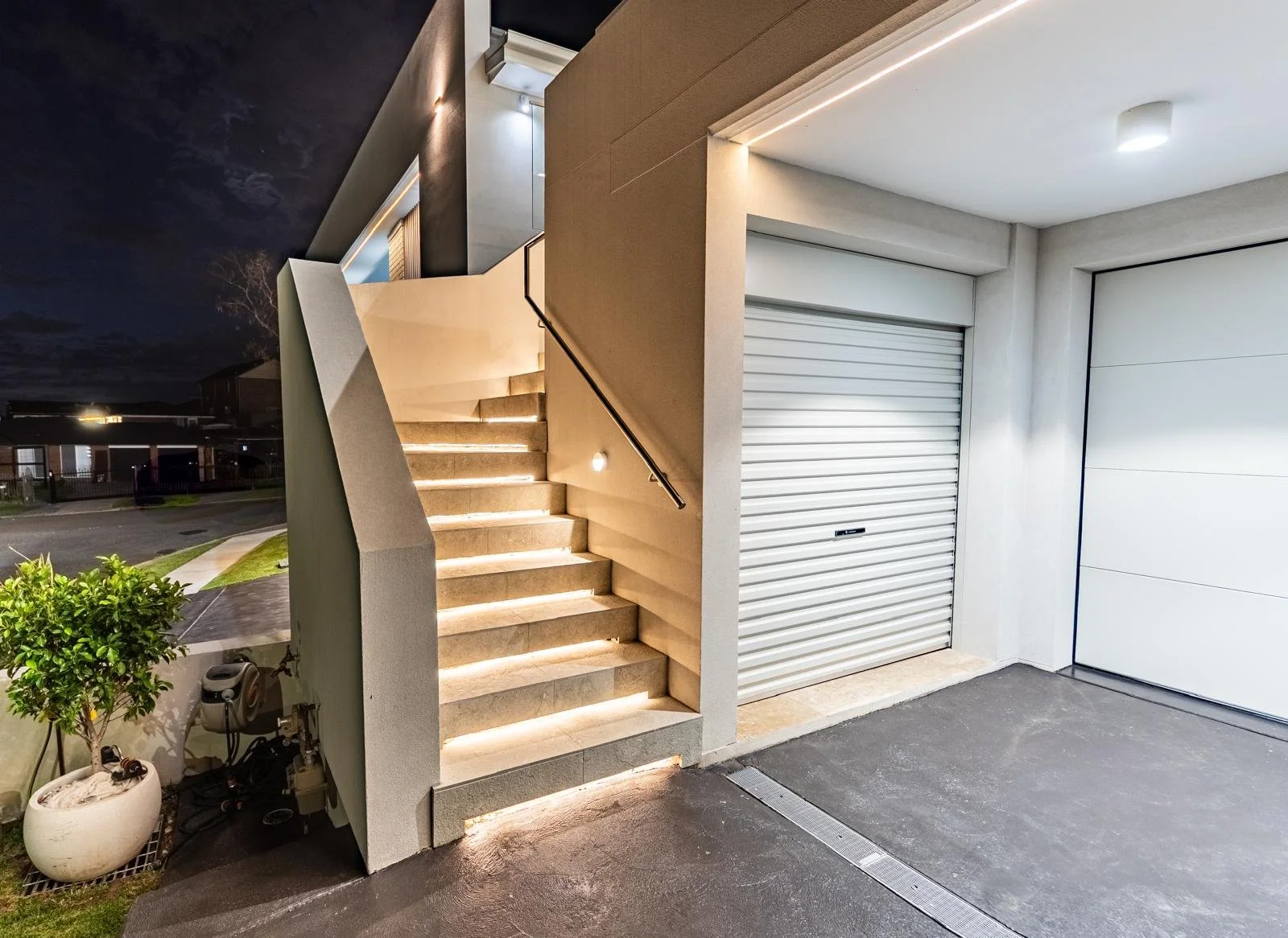 Modern exterior staircase with built-in lighting leading up to a second floor, adjacent to a garage with white roll-up door, in a contemporary residential building at night.