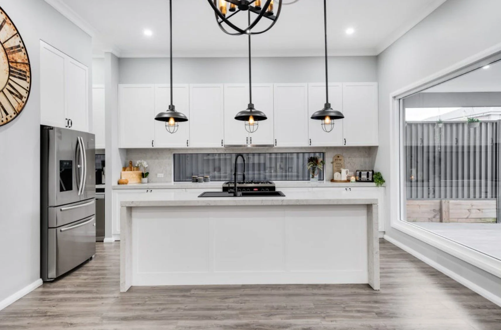 Modern white kitchen with central island, black pendant lights, stainless steel refrigerator, and large sliding glass door leading outside.