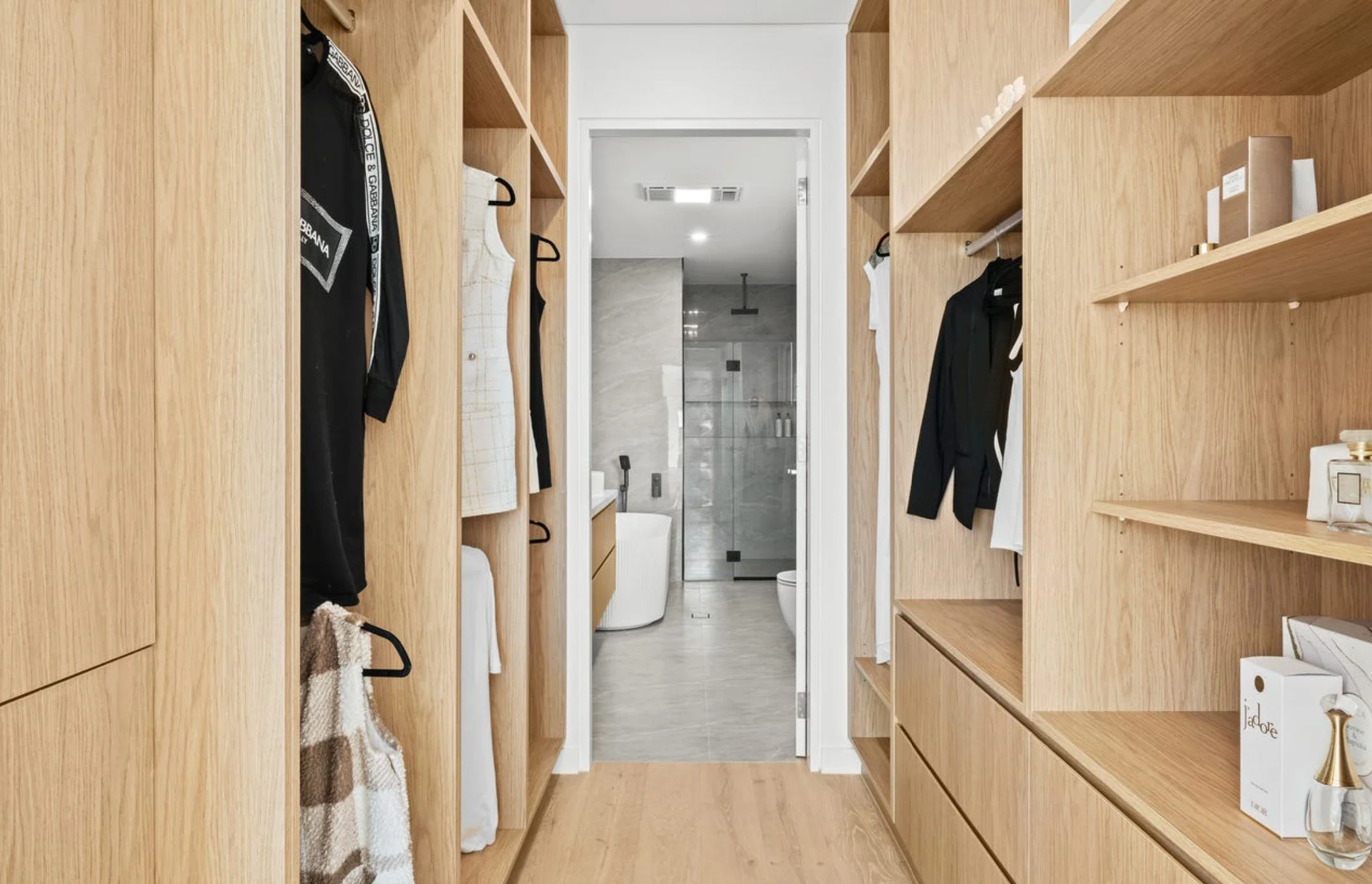 Wooden walk-in closet with shelves and hanging clothes, leading to a bathroom with a gray tiled shower area, bathtub, and toilet.