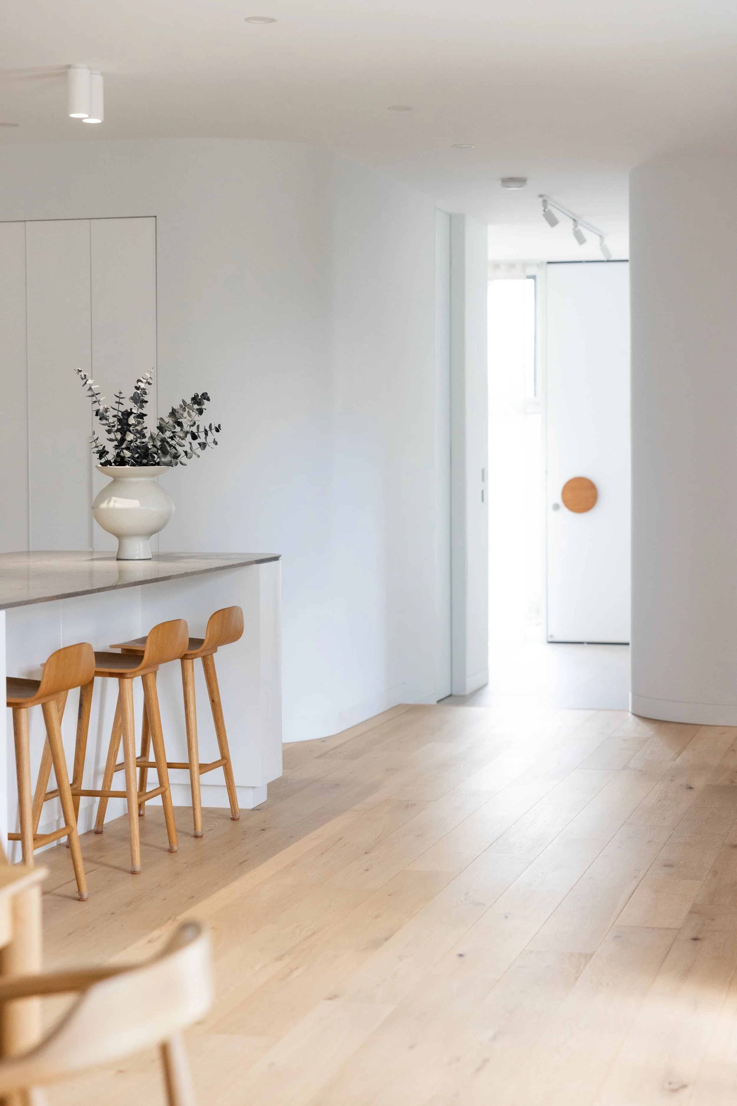 Minimalist kitchen with light wood flooring, white walls, a white vase with black and white eucalyptus on a counter, and three wooden bar stools.