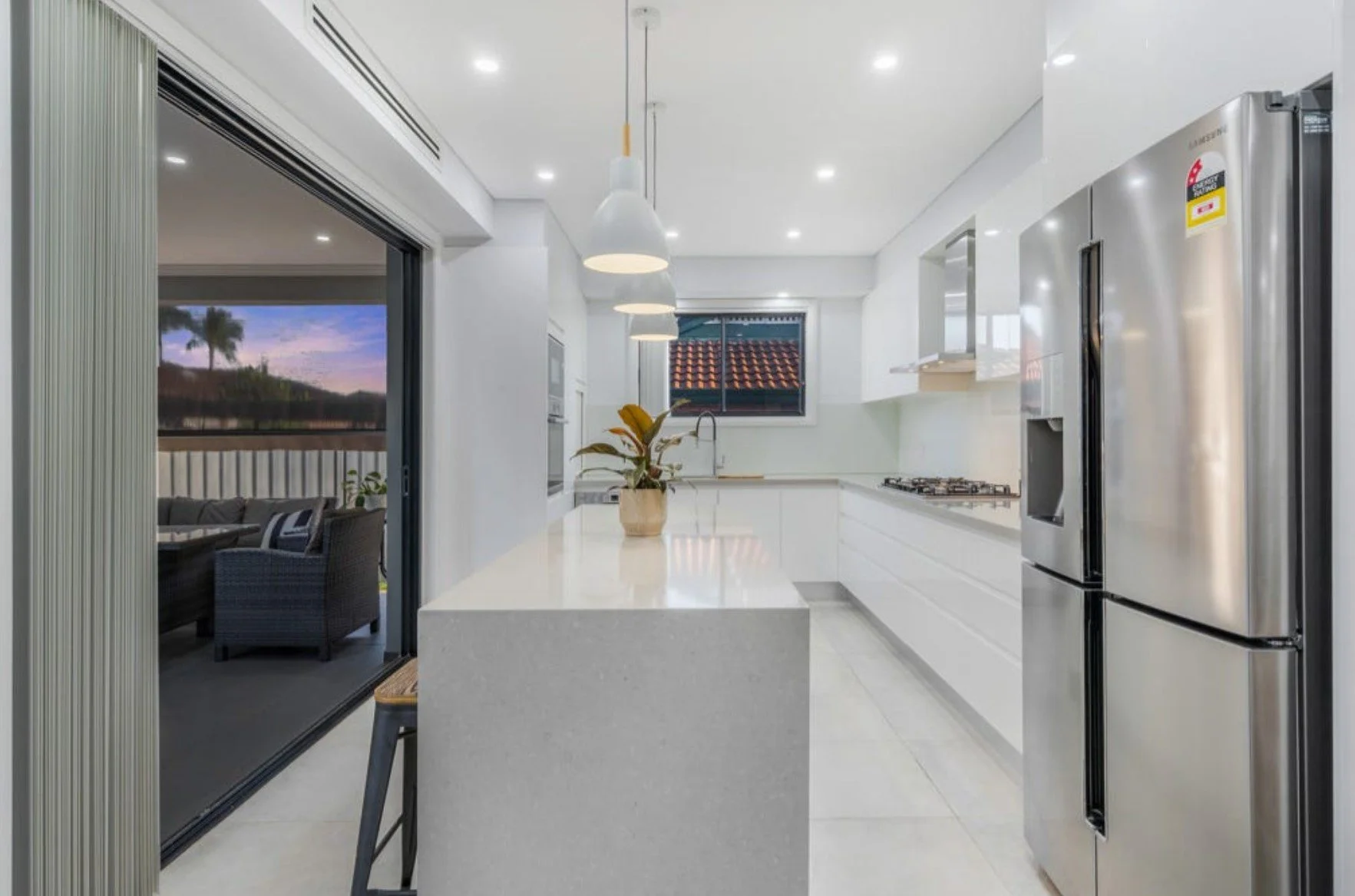 Modern kitchen with white cabinets, a beige island, stainless steel refrigerator, and a window showing an evening sky with palm trees.