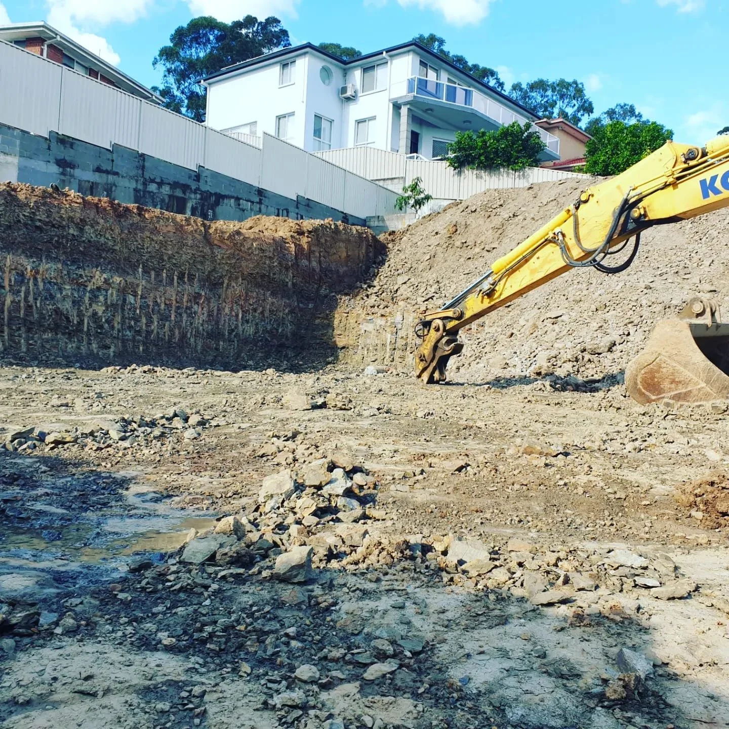 Construction site with excavator digging earth, piles of dirt, and an unfinished area with a backdrop of residential buildings and trees.