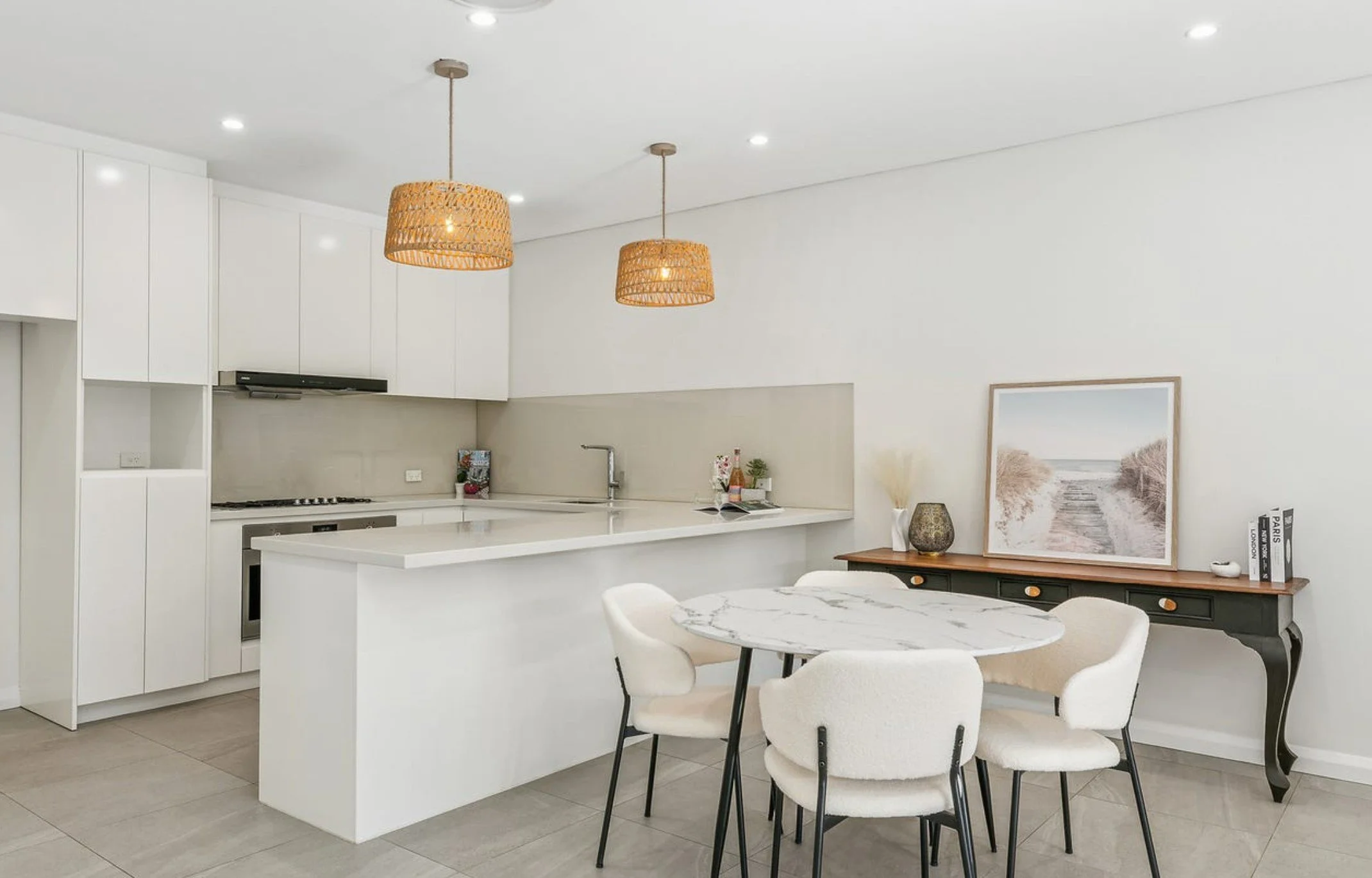 Modern kitchen and dining area with white cabinetry, a marble-topped table, and a decorated sideboard with framed beach artwork.