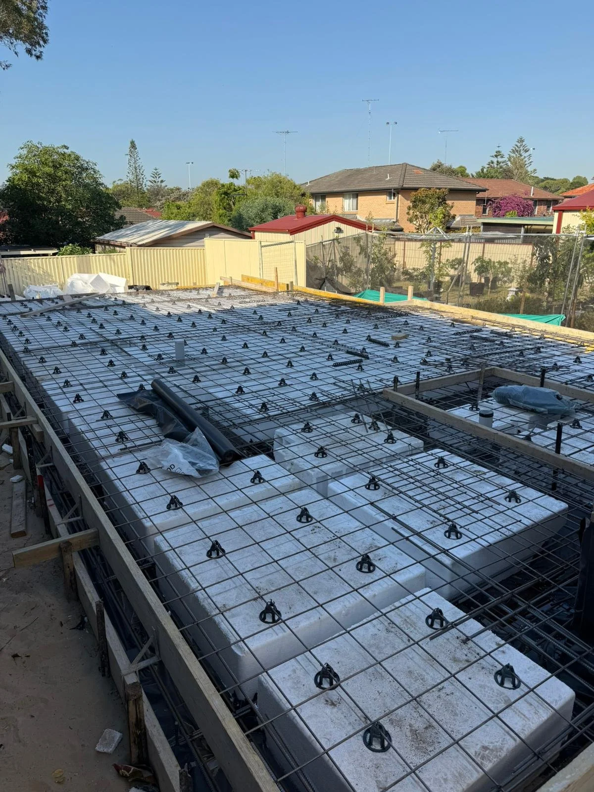 Construction site with steel reinforcement and large white polystyrene blocks on a building foundation, surrounded by fencing and residential houses in the background.
