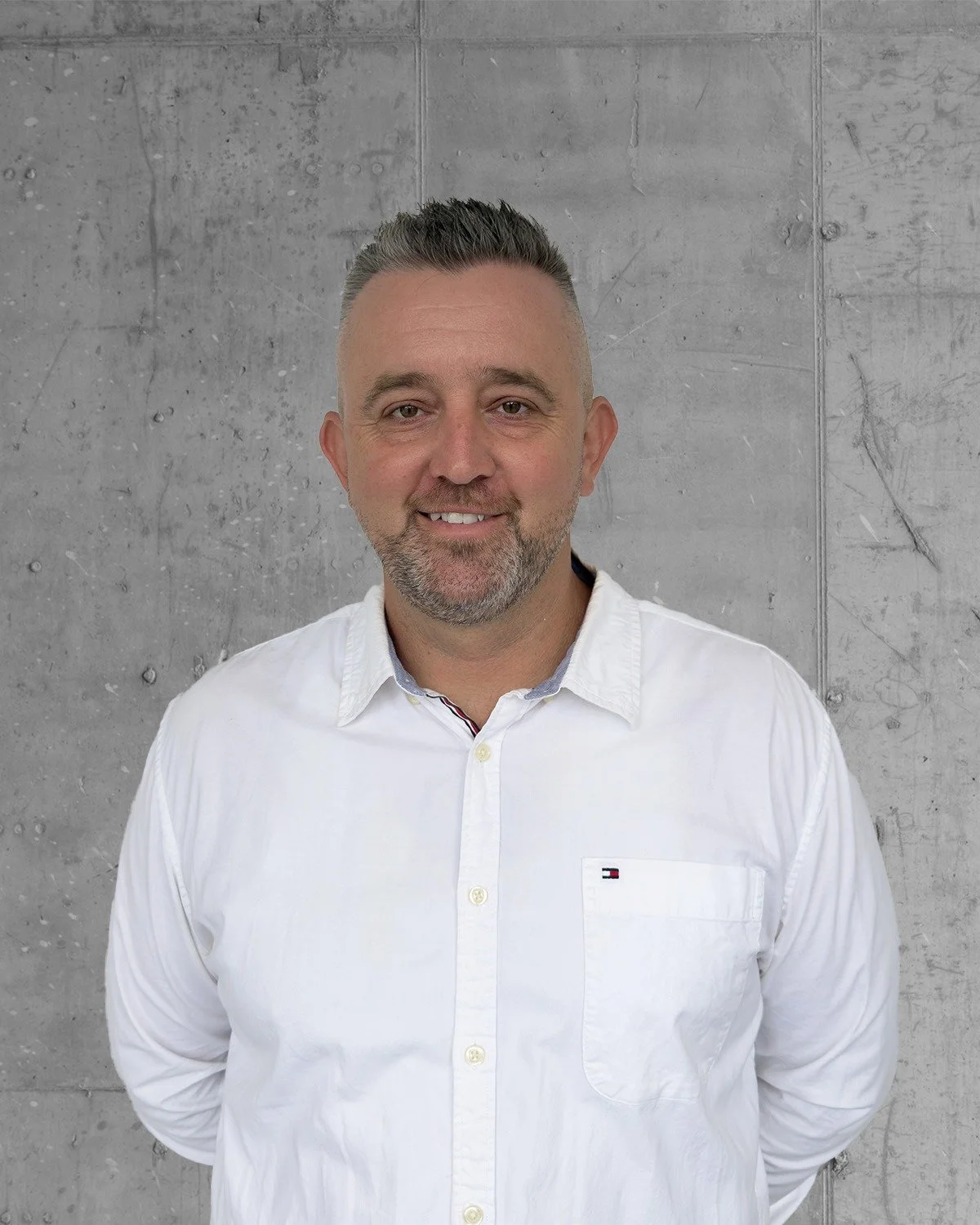 A man with short gray hair and a beard, wearing a white button-up shirt, is smiling and standing in front of a gray concrete wall.