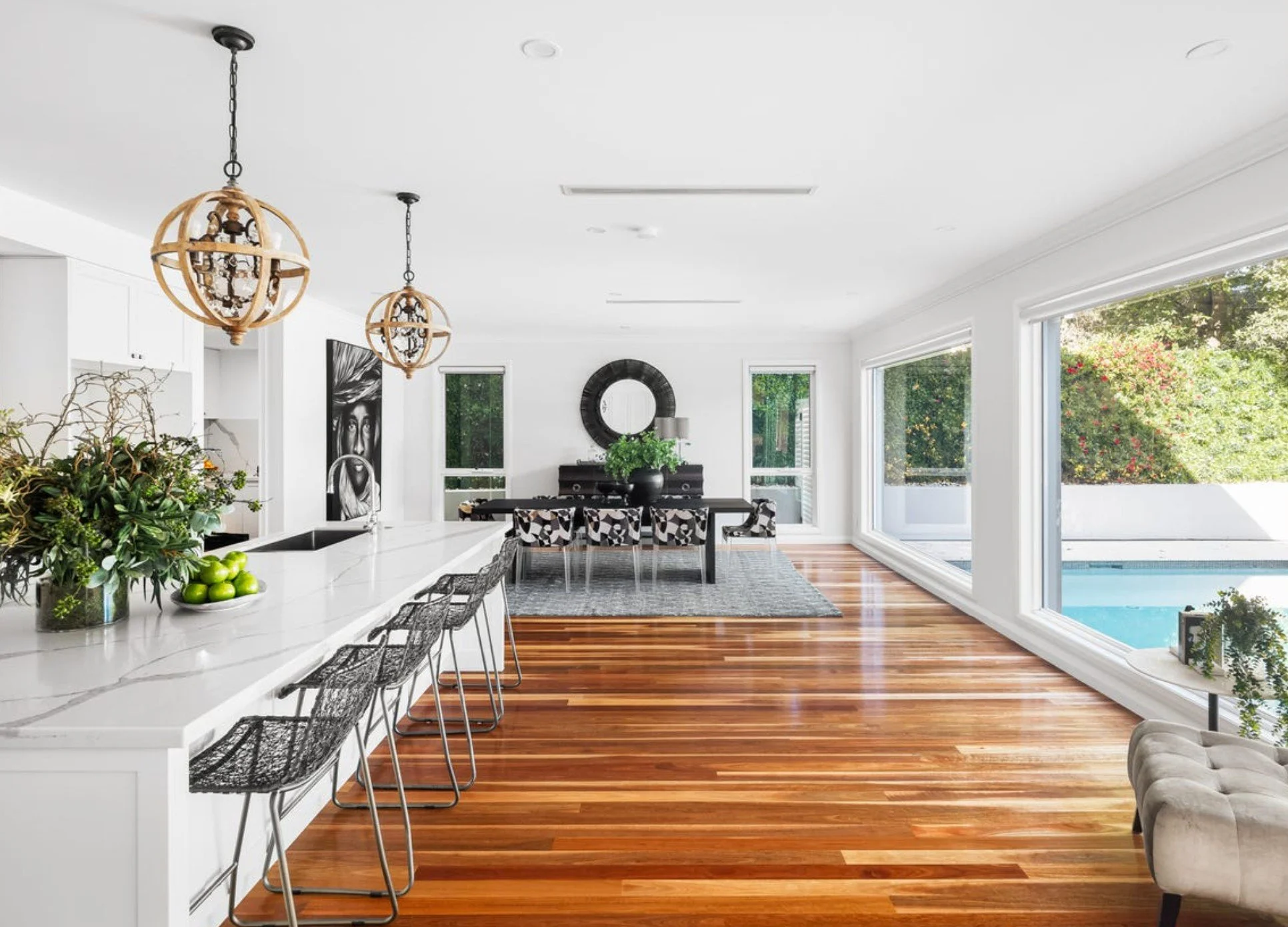 Bright, modern open-concept living space with white walls, hardwood flooring, large windows, a kitchen island with barstools, and a sitting area with a dining table and chairs near the pool outside.