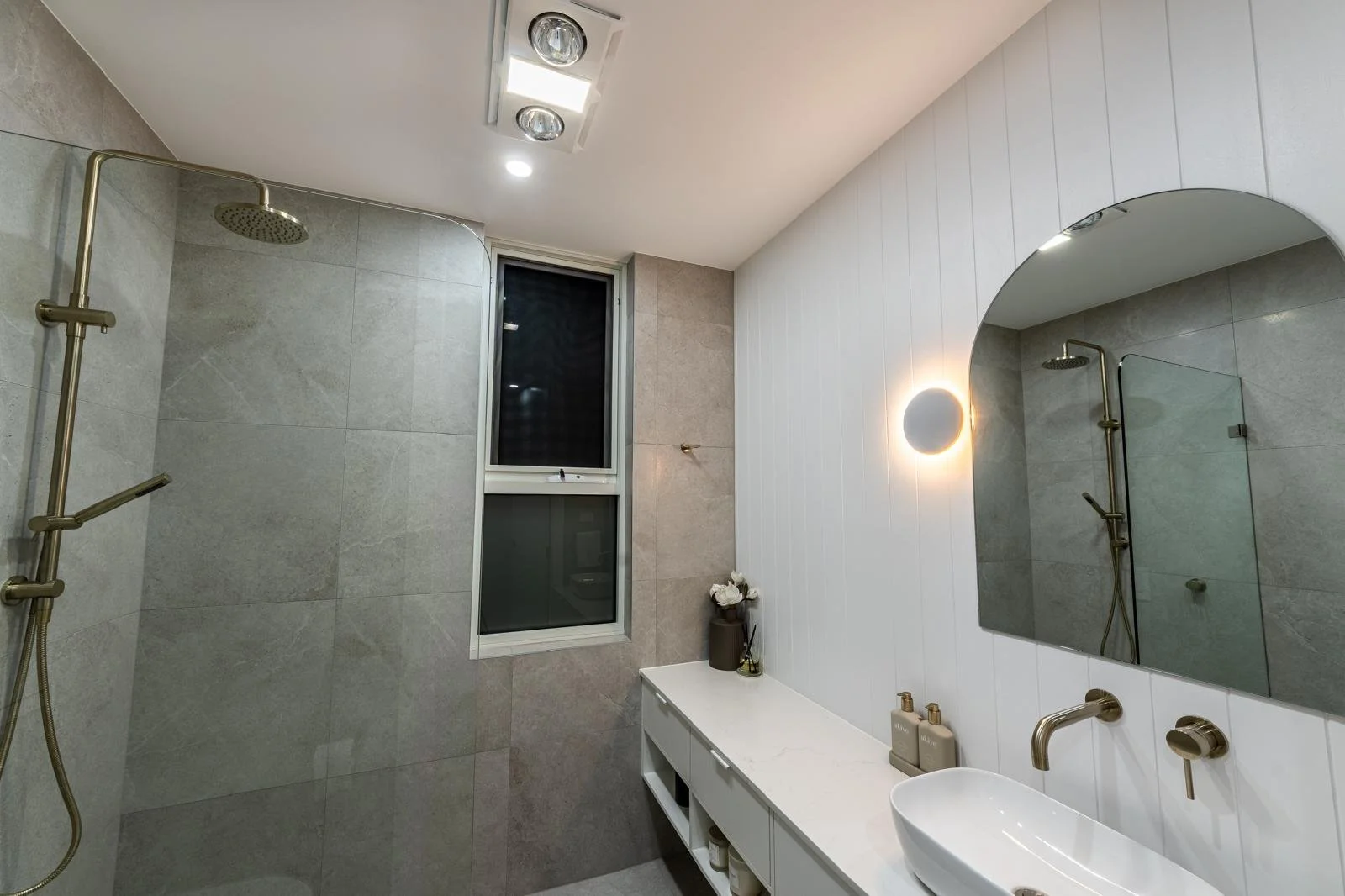 Modern bathroom featuring a walk-in shower with a brass rainfall showerhead, a small window, a large mirror, a white sink on a vanity with decorative bottles, a small round wall-mounted light fixture, and a vase with white flowers.