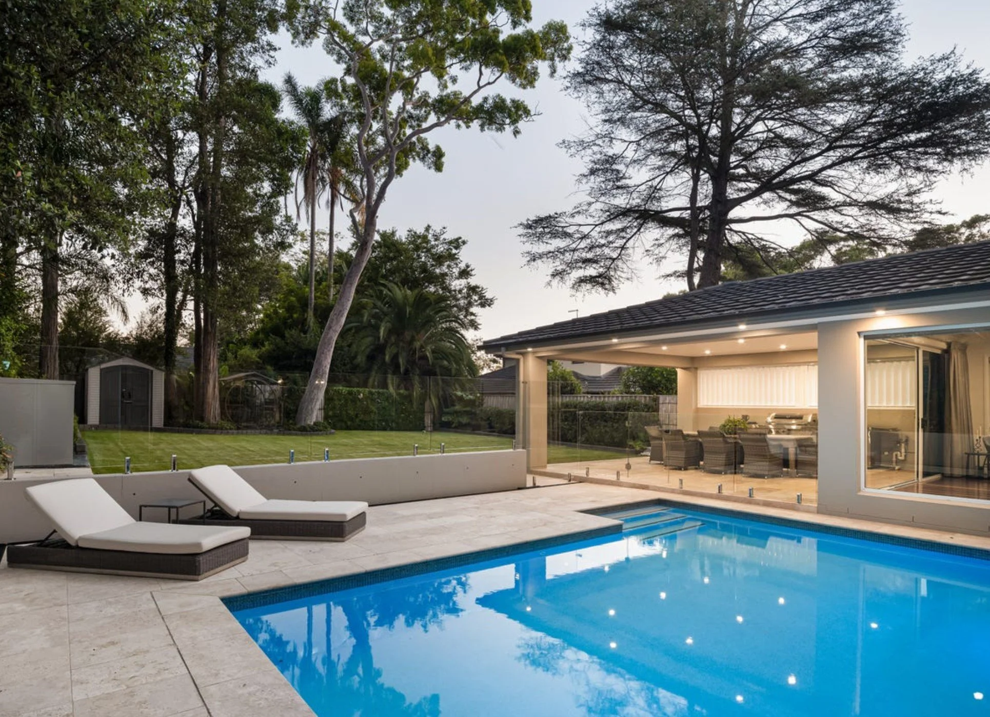 Modern backyard with swimming pool, two lounge chairs with white cushions, glass fence, grassy lawn, tall trees, a covered patio area with outdoor seating, and a house with large windows and sliding door