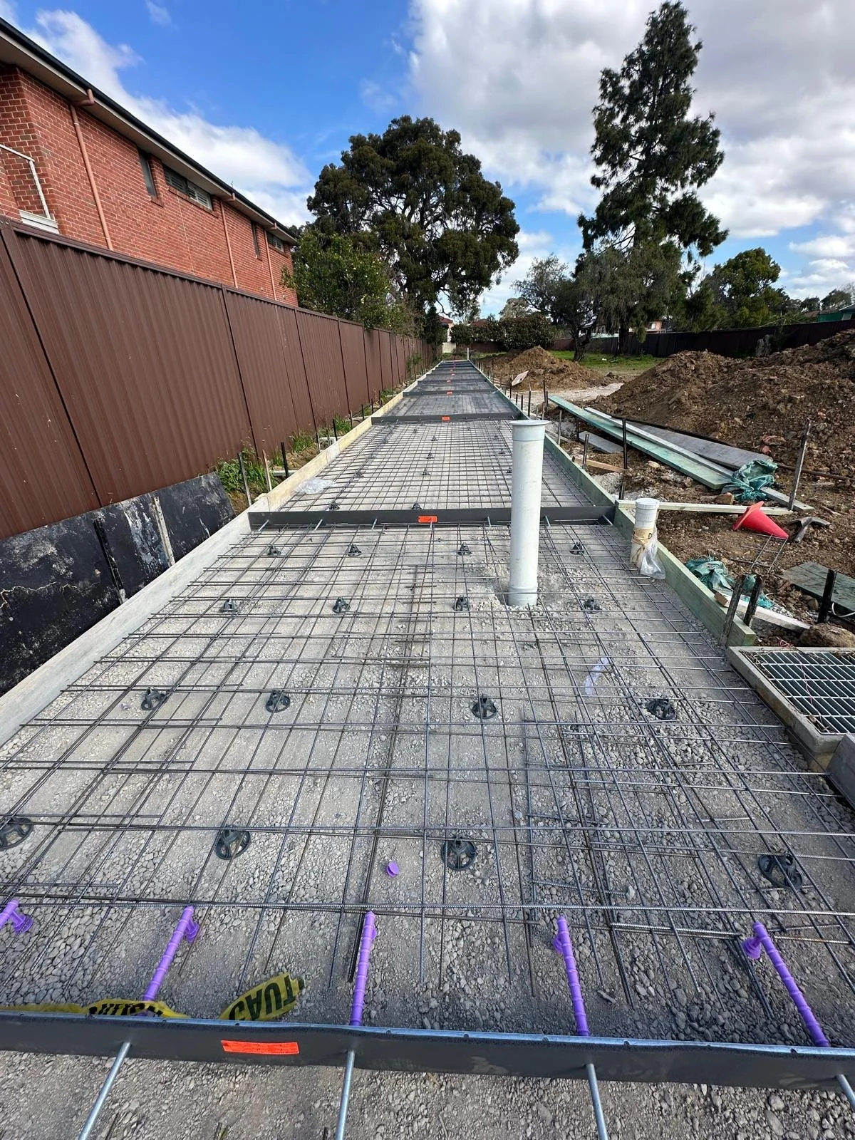 Construction site with rebar framework for a concrete slab, drain pipes, and a view of trees and partially cloudy sky in the background.