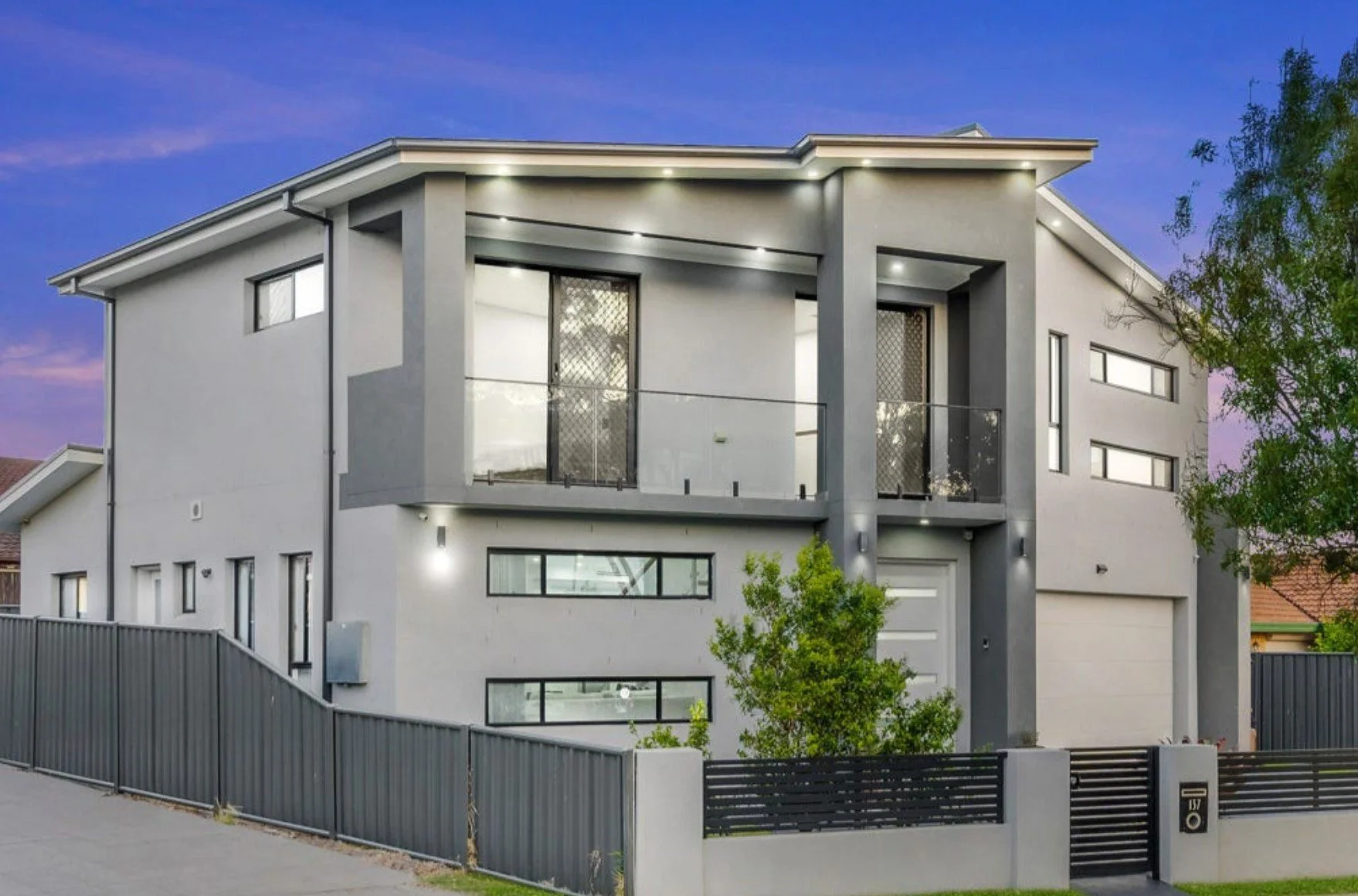 Modern three-story house with gray and white exterior, large windows, a small balcony with glass railing, and a black fence surrounding the property during twilight.