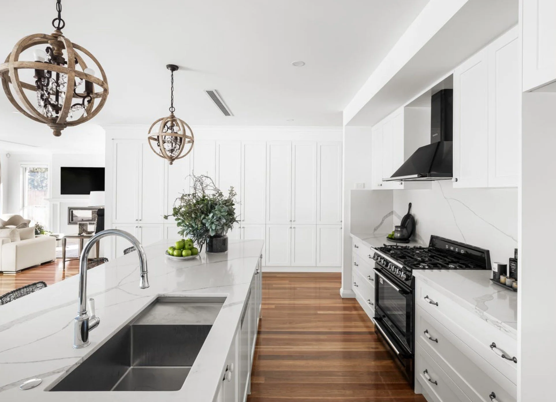 Modern white kitchen with marble countertop island, black range oven, black vent, hanging light fixtures, and hardwood floors, overlooking a living room with a TV.