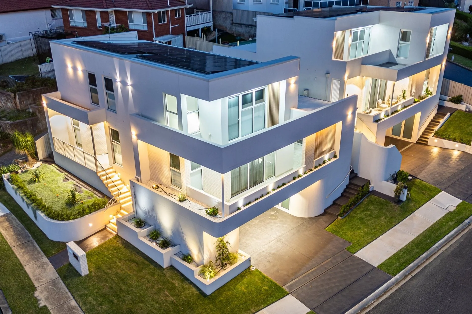A modern multi-story residential building with illuminated exterior lighting, large glass windows, outdoor balconies, stairs, and landscaped yard.