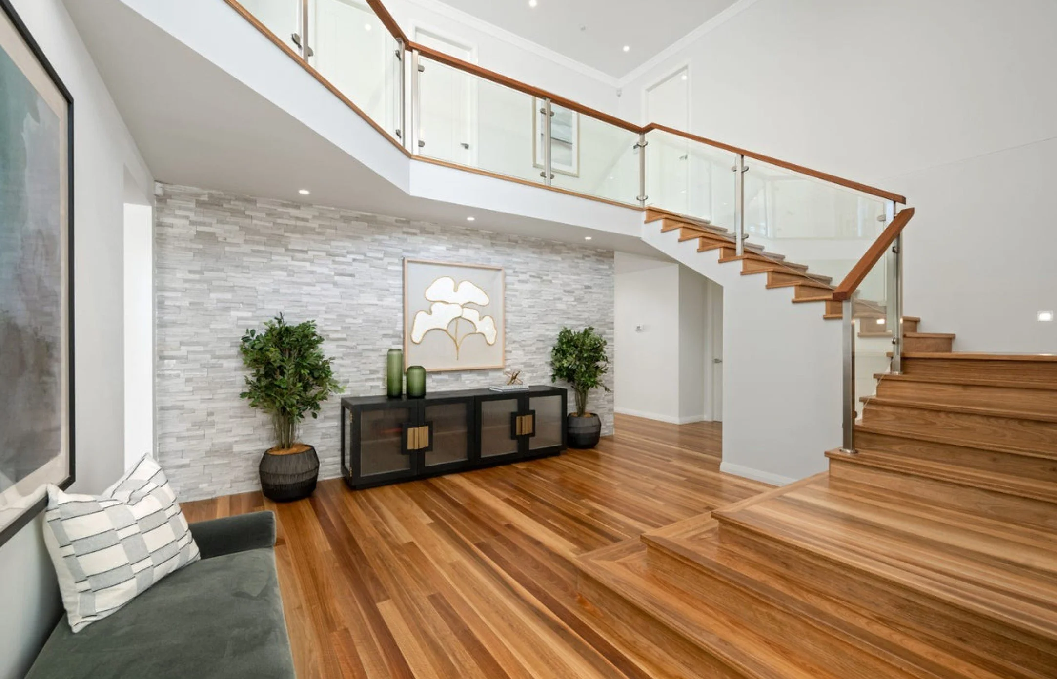 Modern living room with wooden flooring, a staircase with glass and wood railing, a black cabinet with decorative items, twin potted plants, a wall art piece, and a green sofa with striped pillow.