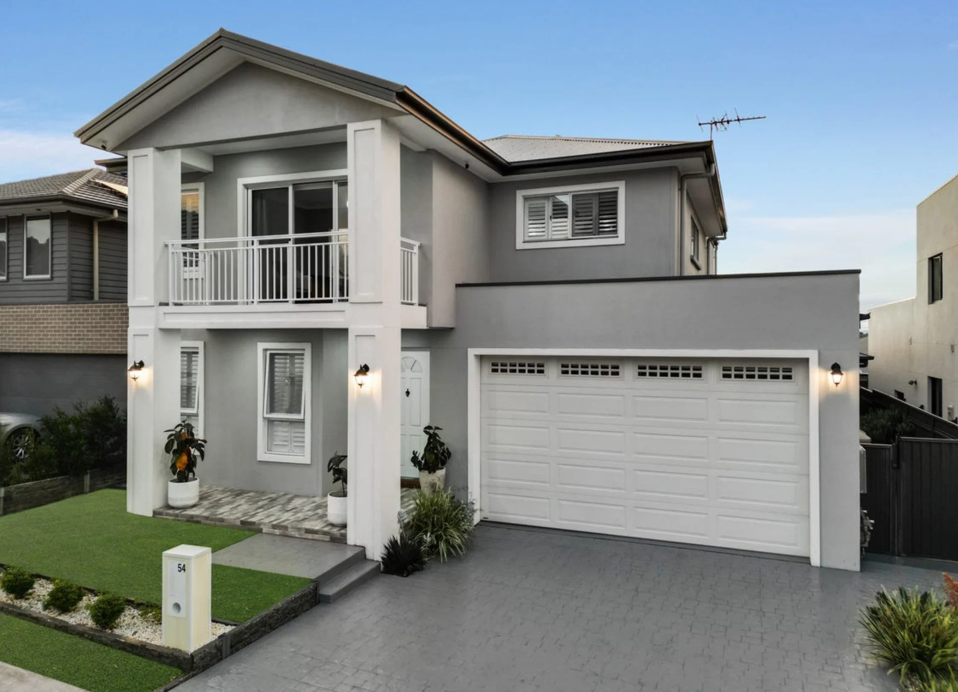 A modern two-story house with a gray exterior, white accents, a front porch with potted plants, and a white garage door. The house has a small front lawn and pathway, with outdoor wall lights and a balcony on the upper floor.