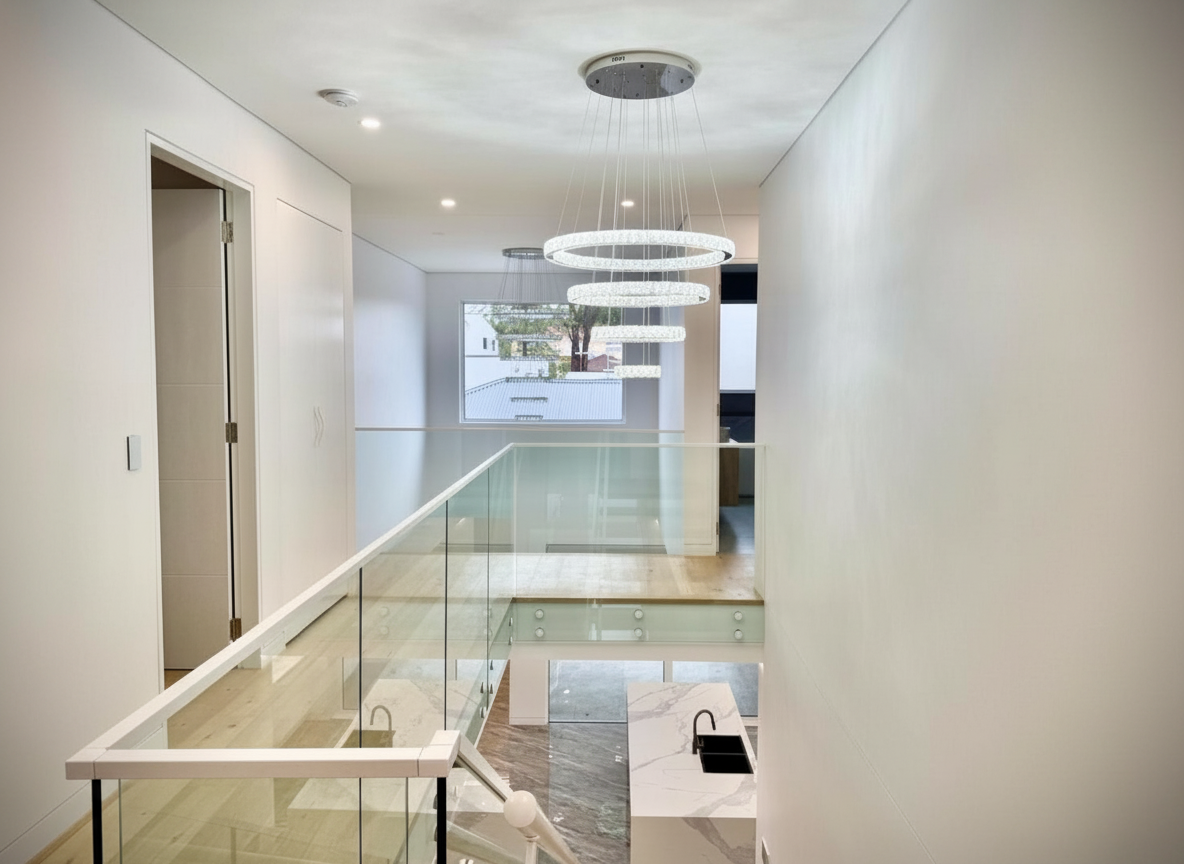 Modern interior with sleek white walls, a floating staircase with glass railing, a contemporary chandelier with circular LED rings, and a large window showing trees and neighboring buildings.
