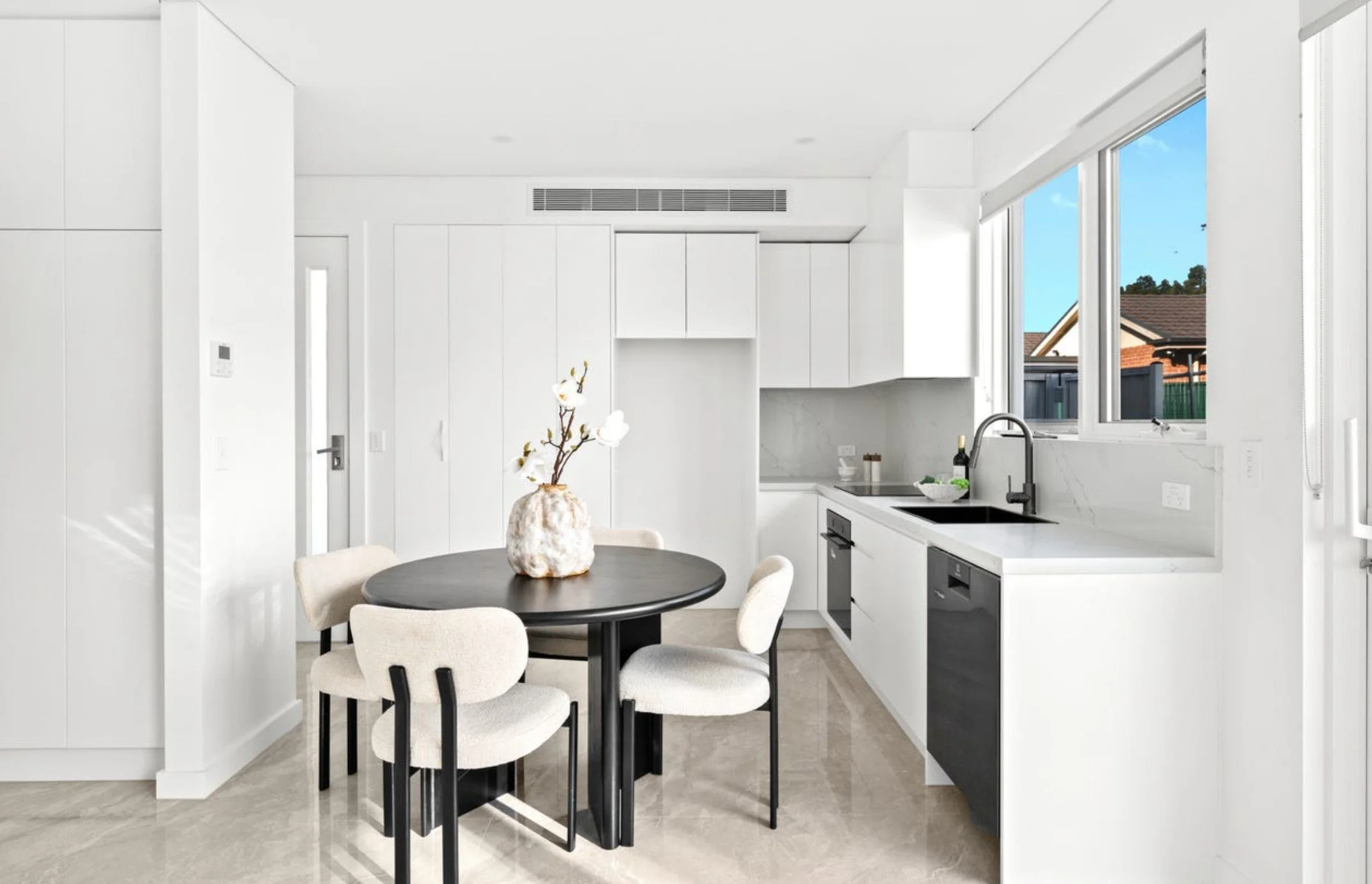 Modern minimalist kitchen with white cabinets, black countertop, and a round dining table with four beige chairs. There are large windows with a view of a neighboring house.