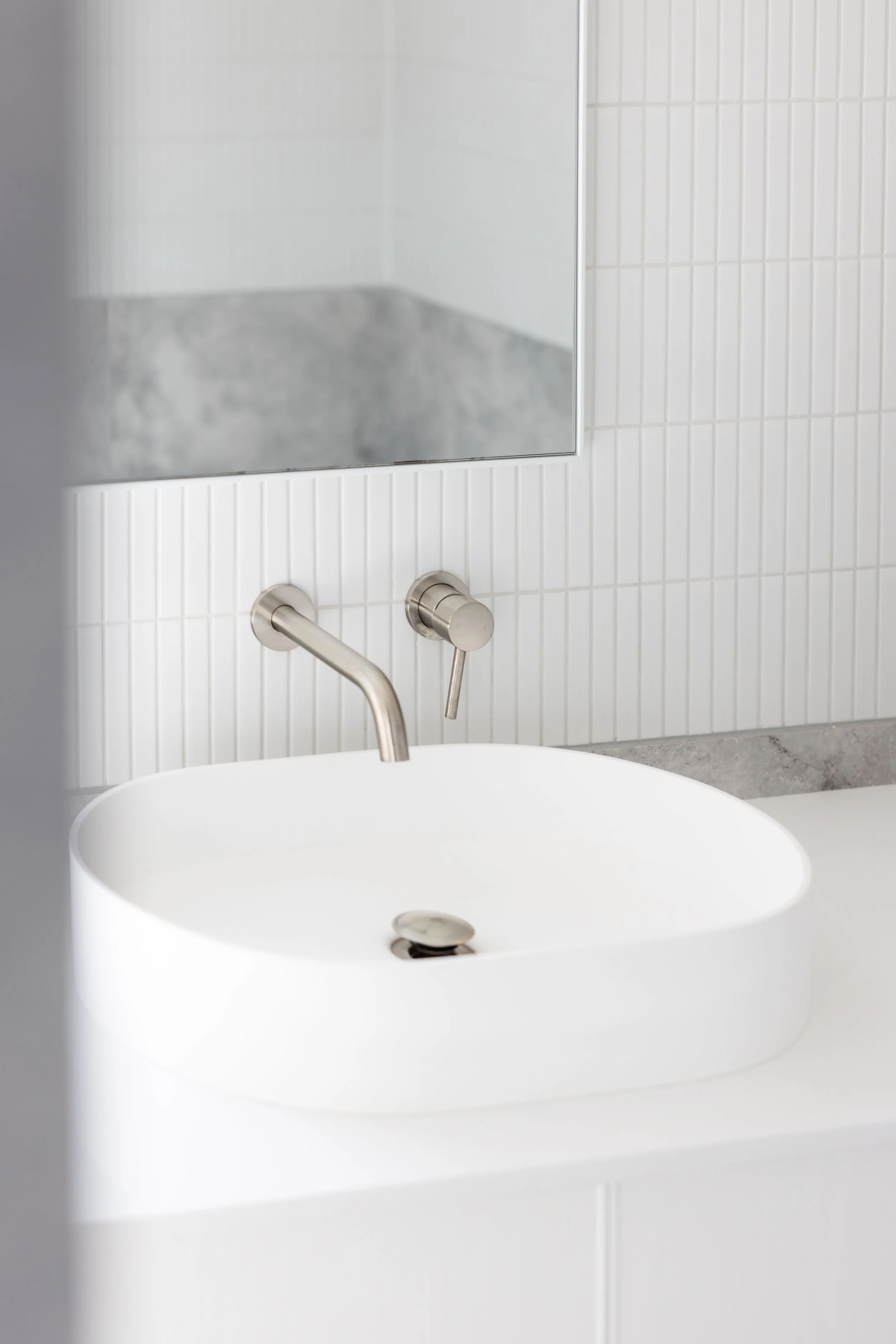 Close-up of a white bathroom sink with a wall-mounted stainless-steel faucet, mirrored backsplash, and white tiled wall.