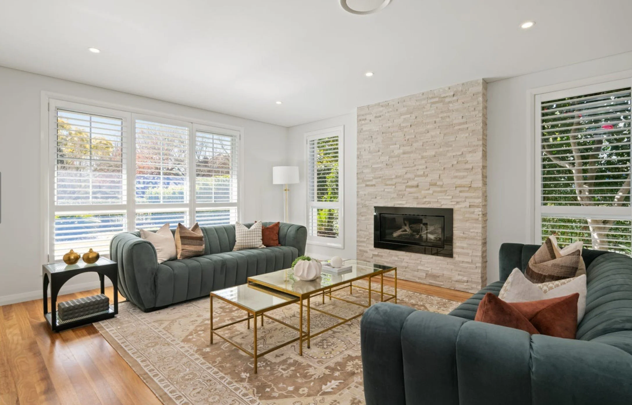Living room with two green sofas, a glass coffee table with gold frame, window with white blinds, fireplace with stacked stone surround, area rug, side table with decorative vases, and a floor lamp.