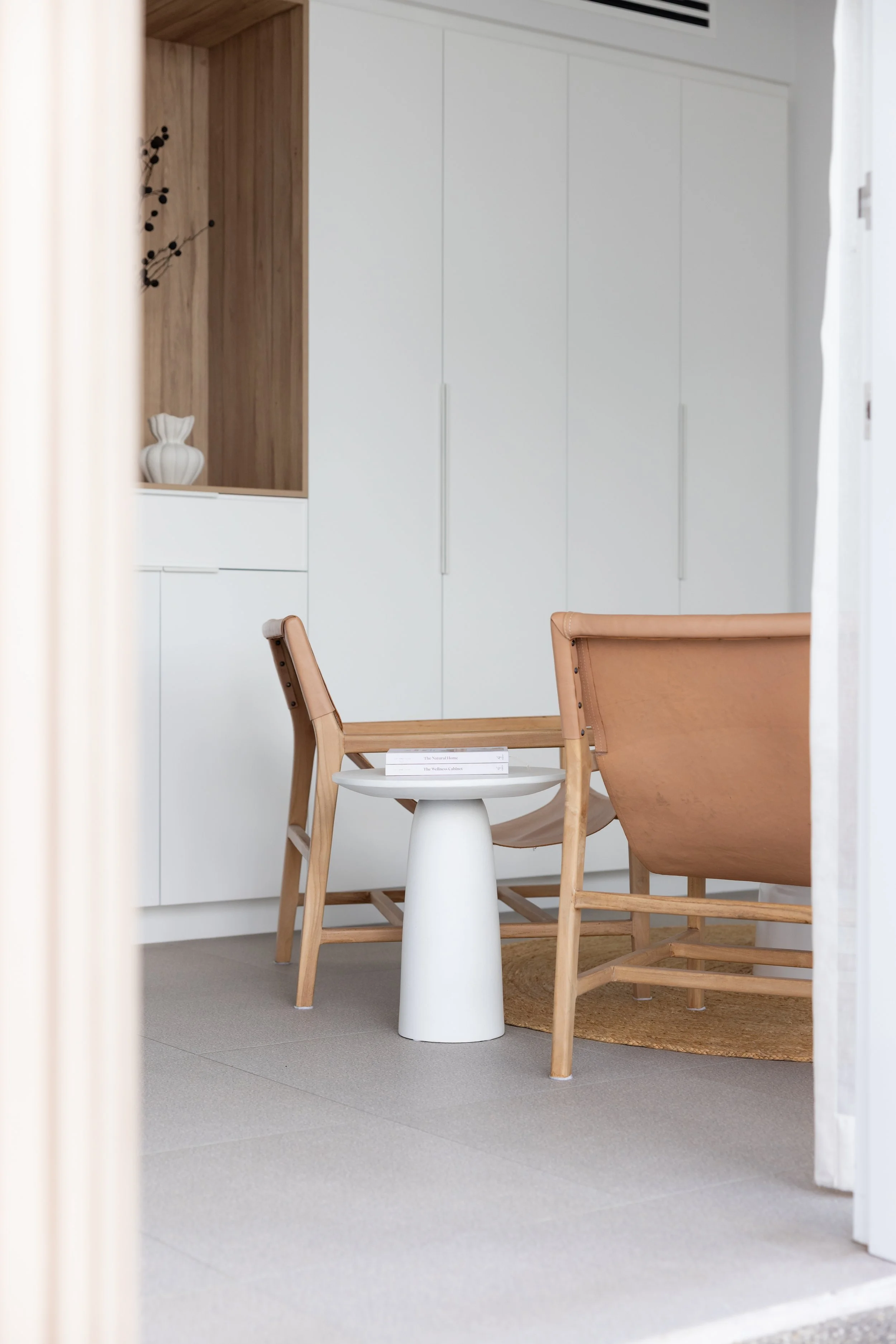 Interior view of a modern room with a white wardrobe, wooden chairs, a white side table with books, a woven rug, and a minimalist decor.