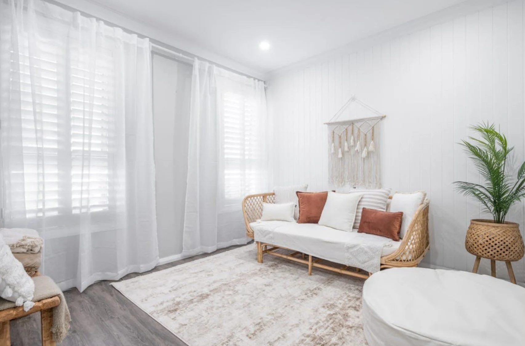 Bright living room with white walls, sheer curtains, beige sofa with white and brown pillows, woven side table, potted plant, plush rug, and wall hanging.