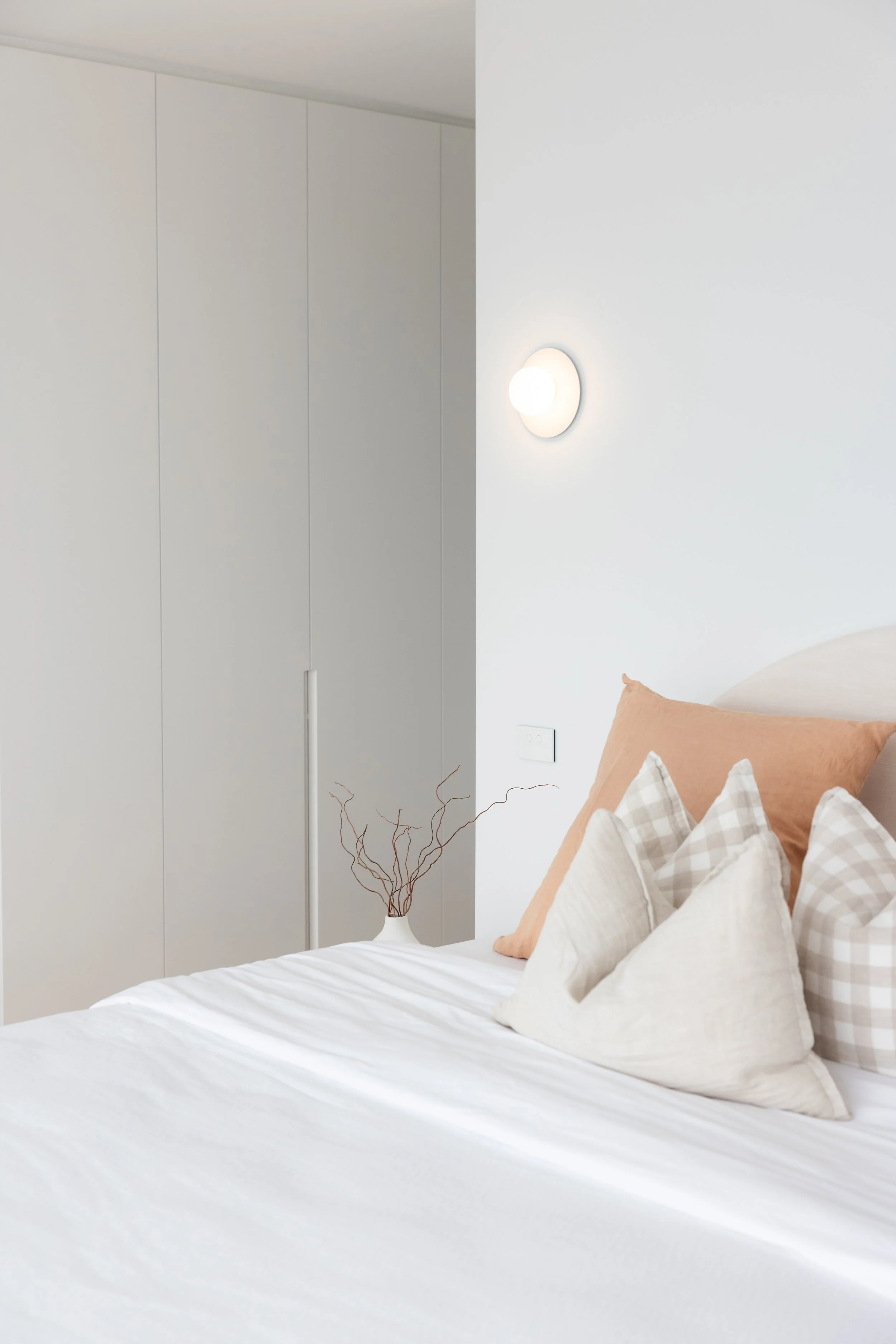 A neatly made bed with beige and plaid pillows in a minimalist white room with a wall-mounted light and a white wardrobe.