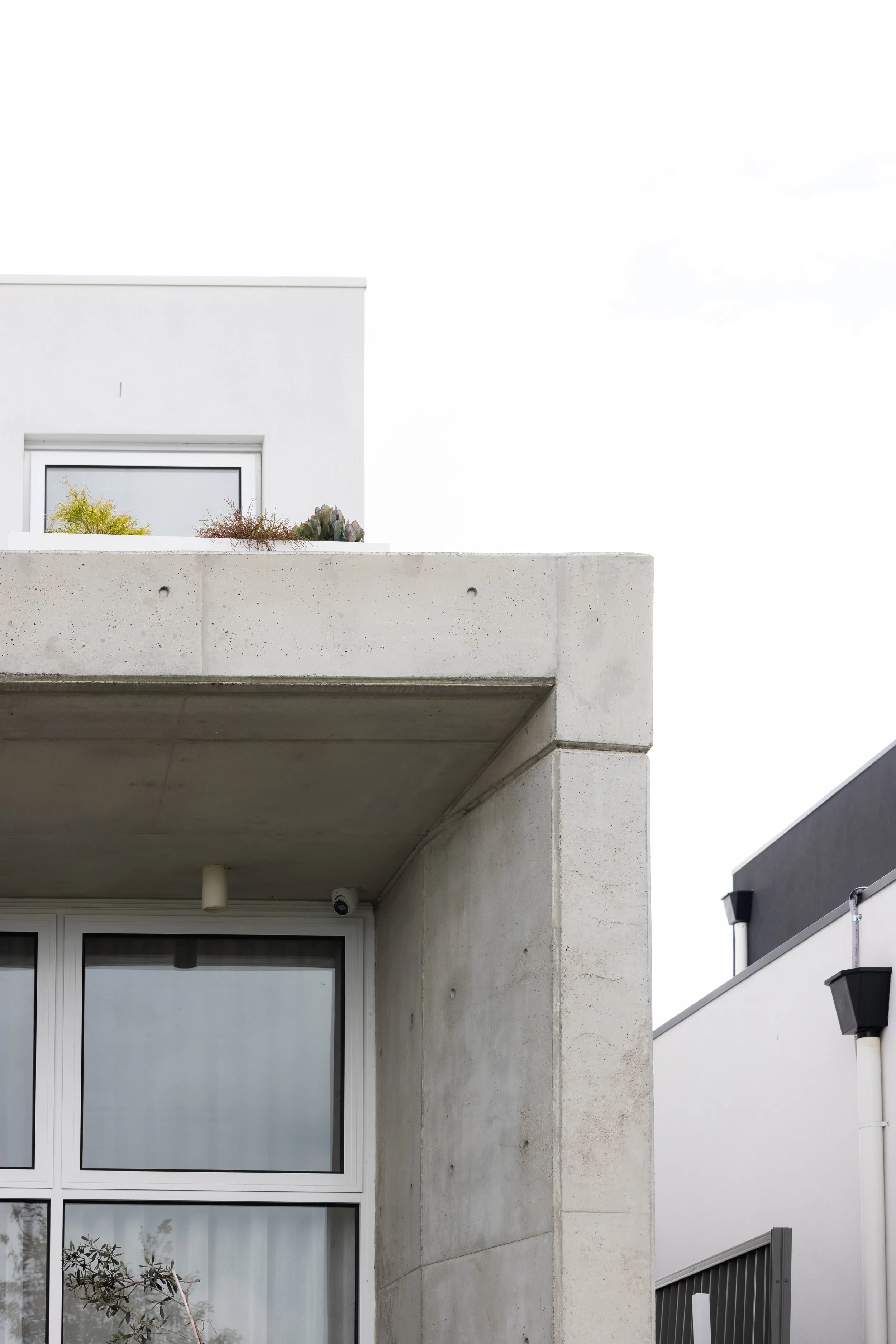 Modern building with concrete and white exterior, large window, small plants on balcony, overcast sky.
