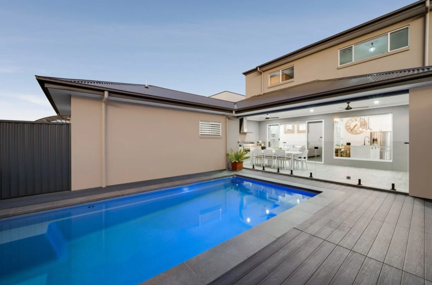 Modern backyard with a swimming pool and an open-plan indoor dining and kitchen area visible through glass sliding doors, featuring a large wall clock and a stainless steel refrigerator.