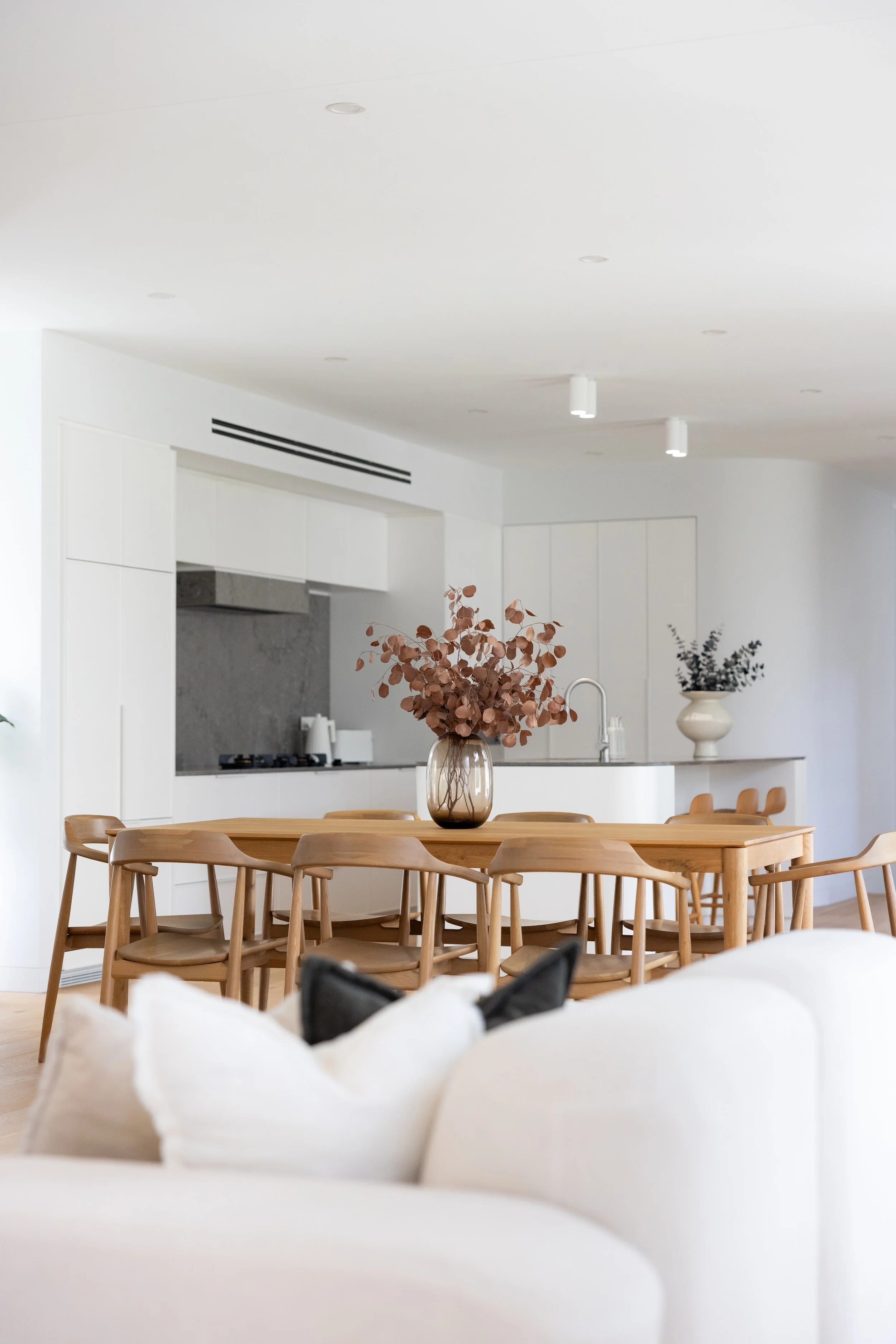 Modern minimalist open-concept living and dining area with a large wooden dining table, surrounding wooden chairs, and a white sofa with cushions in the foreground. The kitchen in the background features white cabinetry, a gray backsplash, and decora