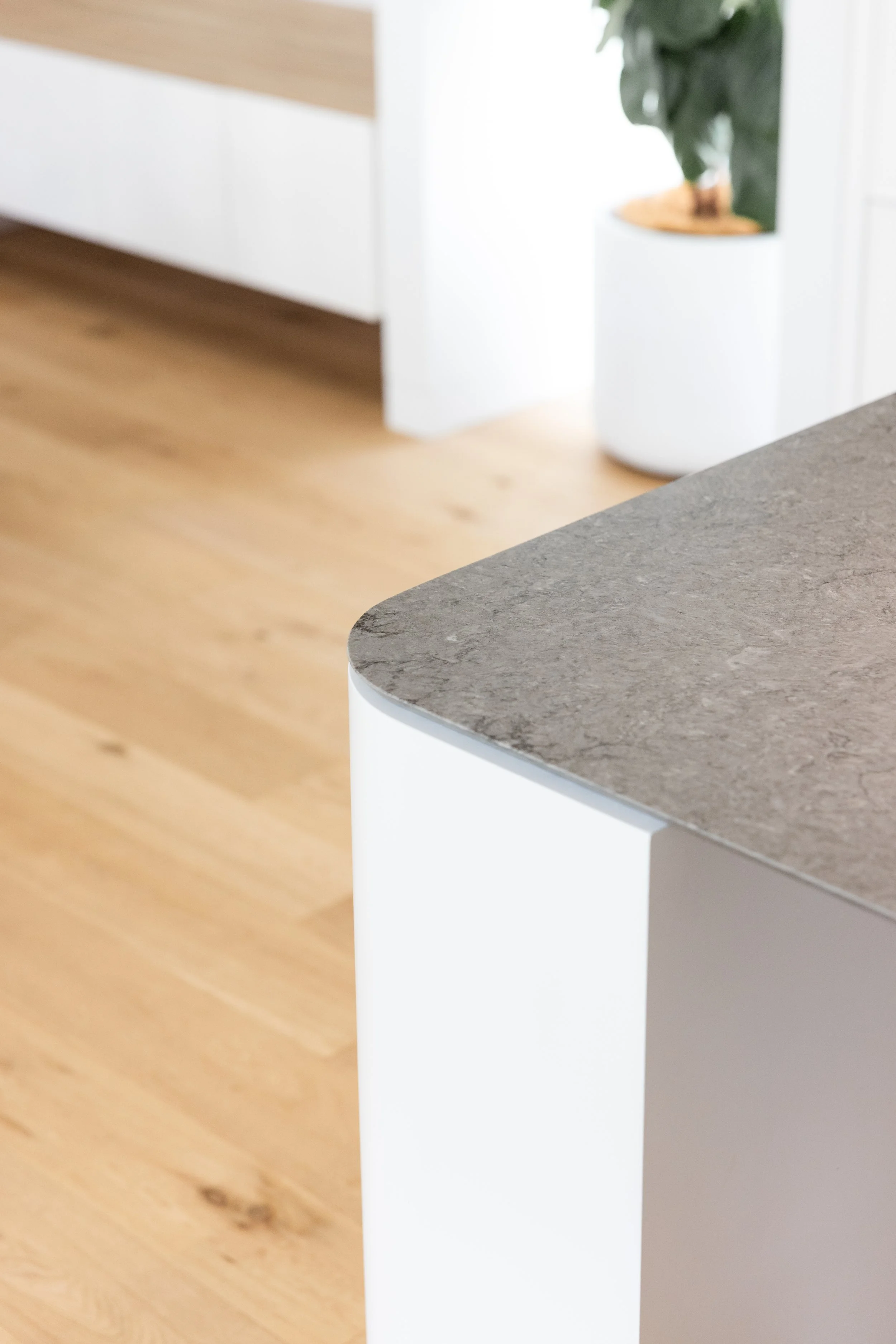 Close-up of a kitchen island with a gray stone countertop and white base, with hardwood flooring and a potted plant in the background.