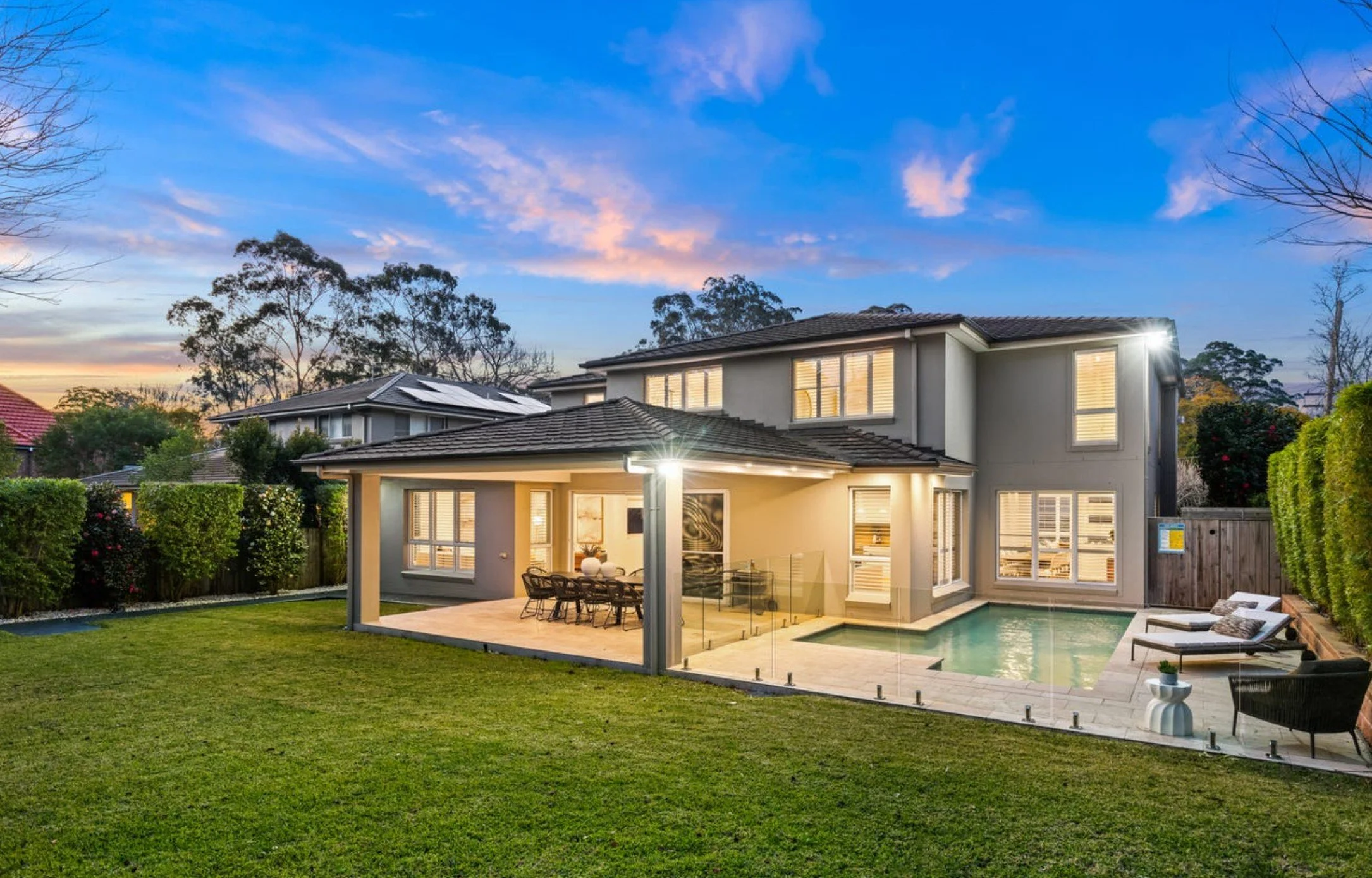 Modern two-story house with backyard pool, outdoor dining area, and lounge chairs, illuminated at dusk