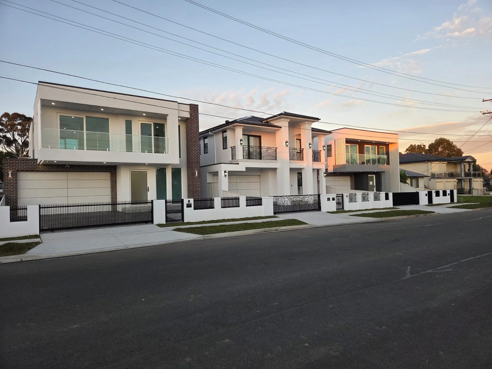 Three modern houses with balconies, gated yards, and a driveway, photographed during sunset.