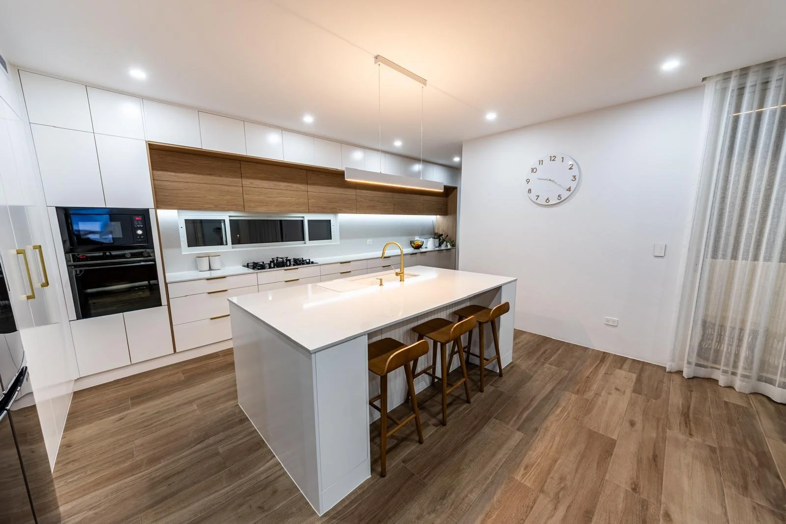Modern kitchen with white cabinets, wooden accents, a central island with a built-in sink and three wooden barstools, a wall clock, and sliding glass doors with sheer curtains.