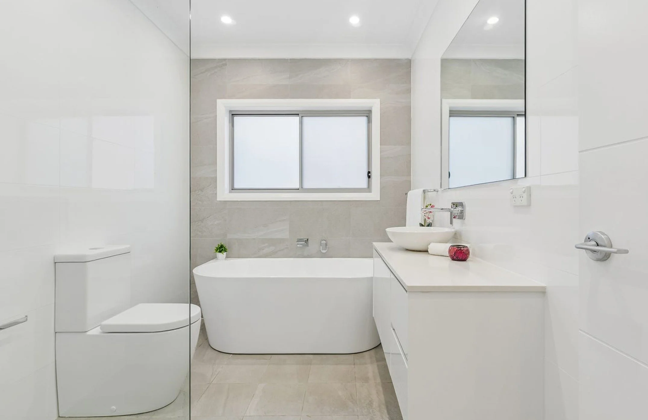 Modern bathroom with white fixtures, a bathtub, a toilet, a large mirror, a window, and minimal decor.