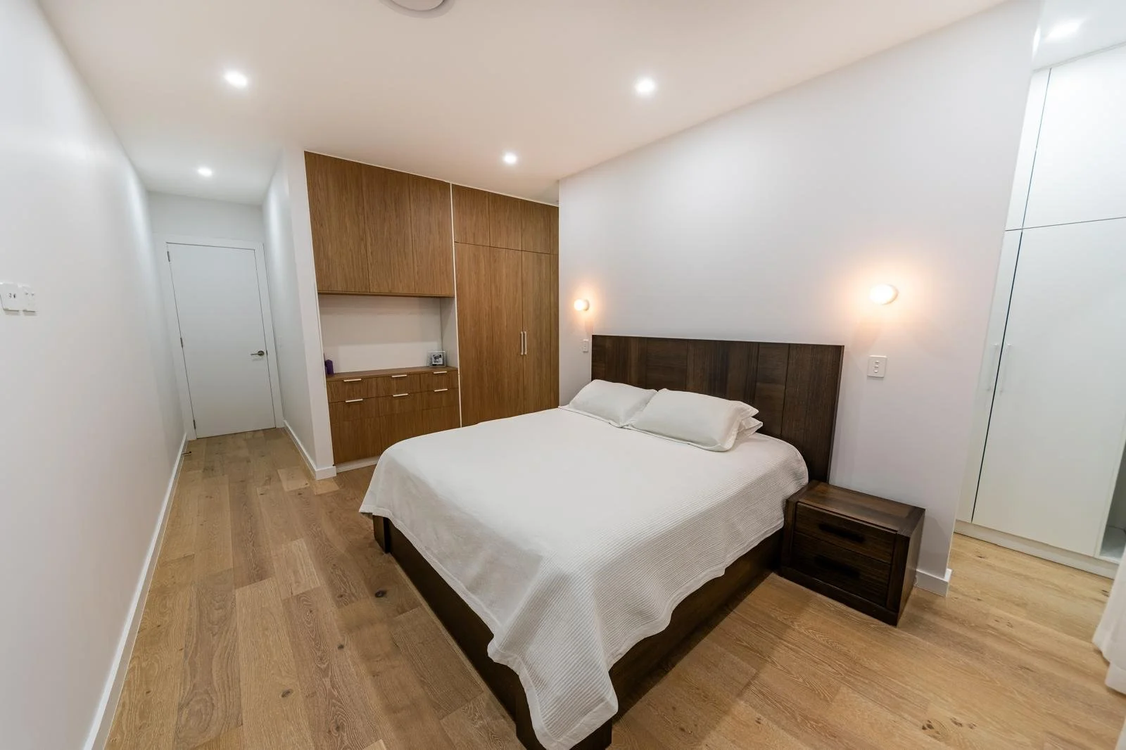Modern bedroom with white walls, wooden flooring, a large bed with white bedding and pillows, dark wood headboard, bedside table, and built-in closet with wooden cabinets, illuminated by ceiling and wall lights.