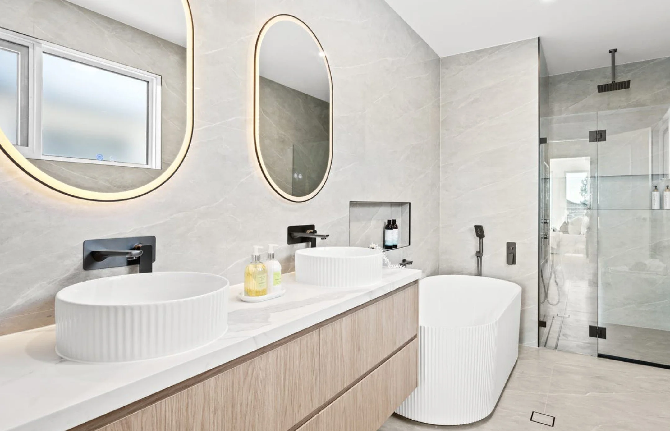 Modern bathroom with double oval vessel sinks, wall-mounted black faucets, wood vanity, large backlit mirrors, a bathtub, and a glass-enclosed shower with rainfall and handheld showerheads.