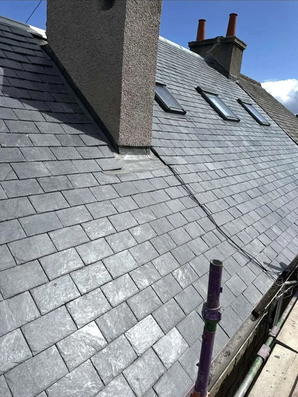 A rooftop with gray slate shingles, three skylight windows, and a chimney with two red chimney pots.