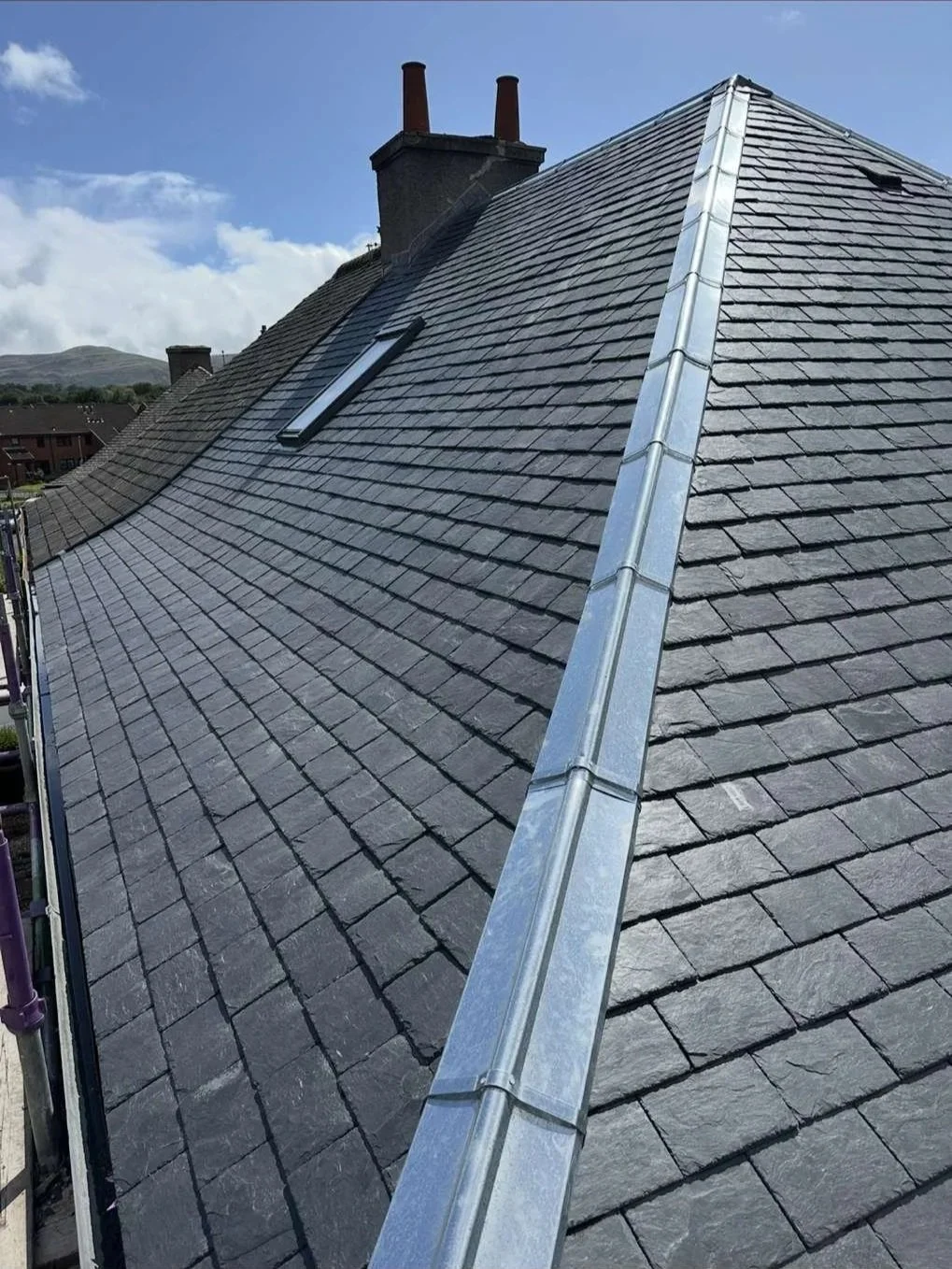 Roof with slate shingles, a skylight, and metal flashing along the ridge, with chimneys visible in the background.
