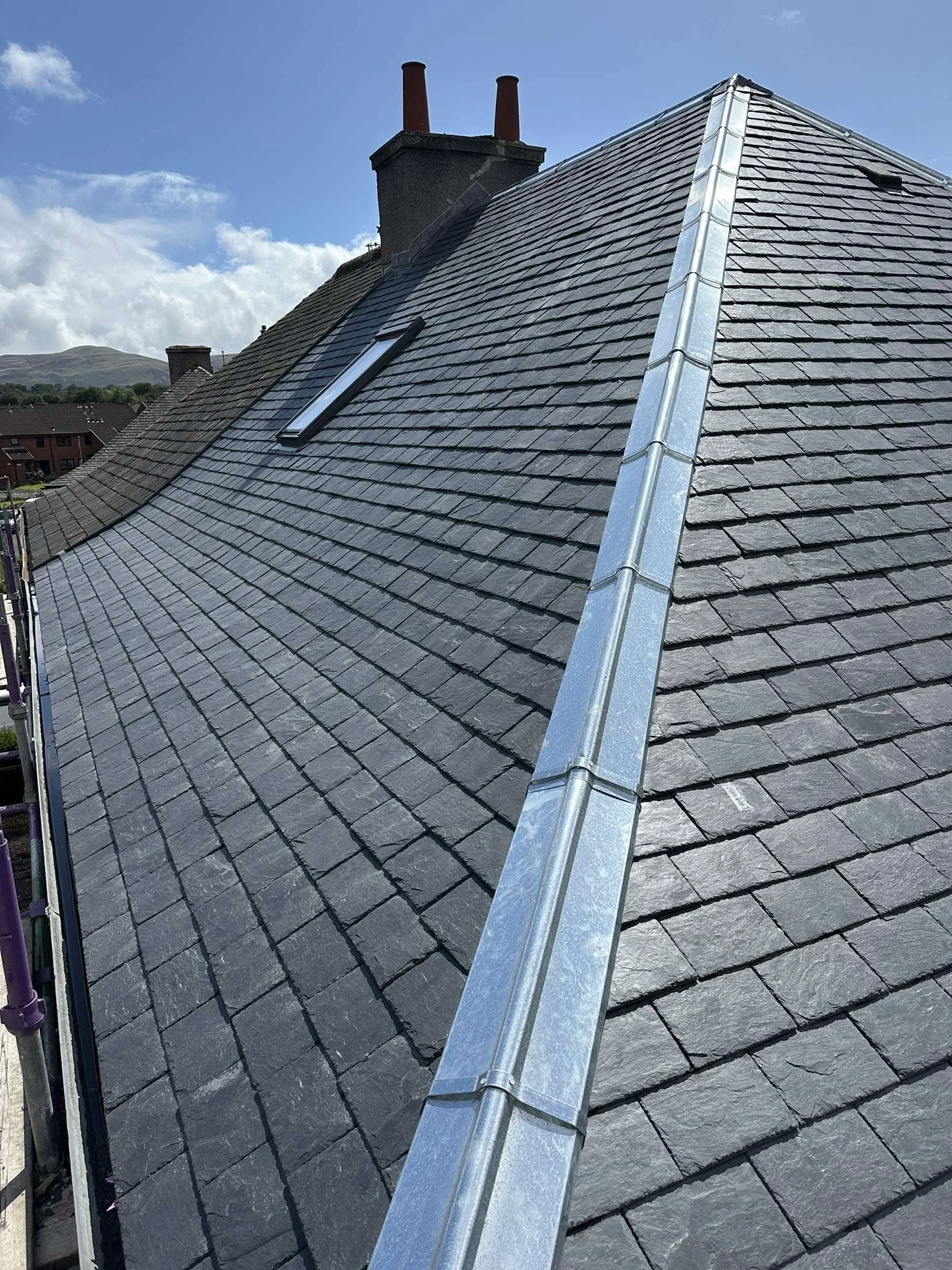 View of a house roof with dark gray slate tiles, skylight window, and metal flashing along the ridge under a partly cloudy sky with mountains in the background.