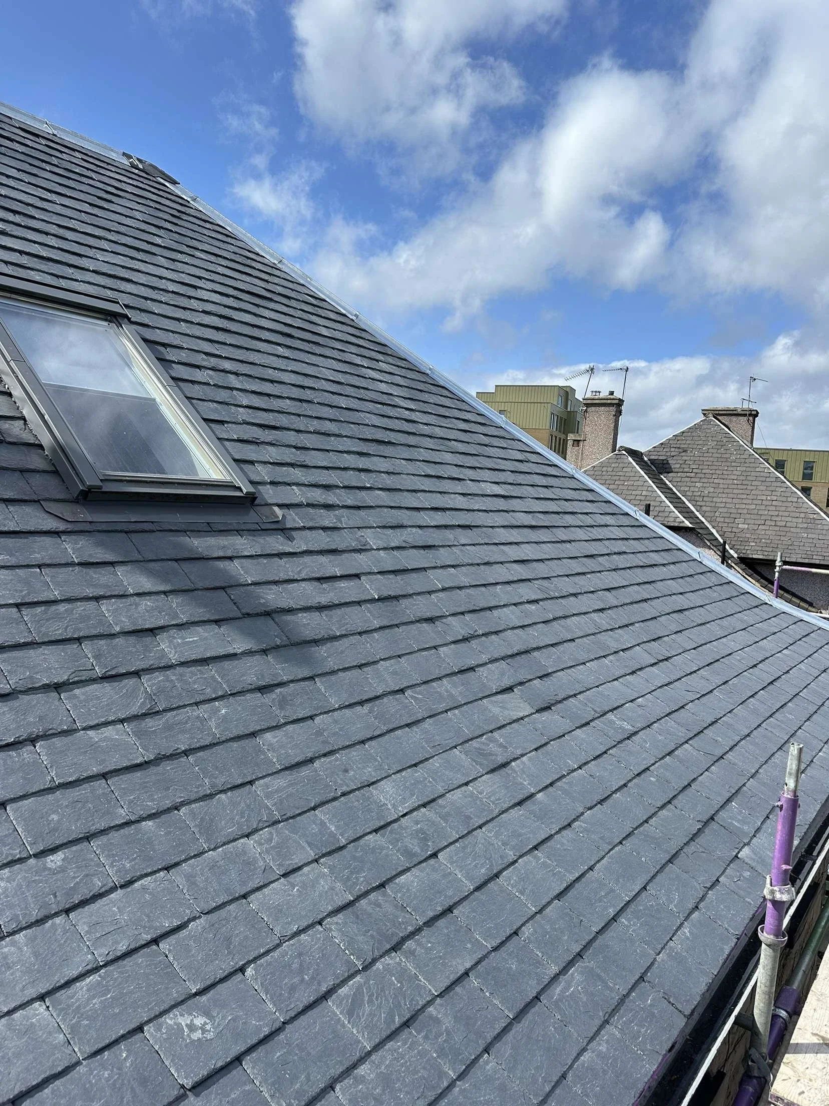 Close-up view of a dark gray slate tiled roof with a skylight window, under a partly cloudy blue sky with neighboring rooftops in the background.