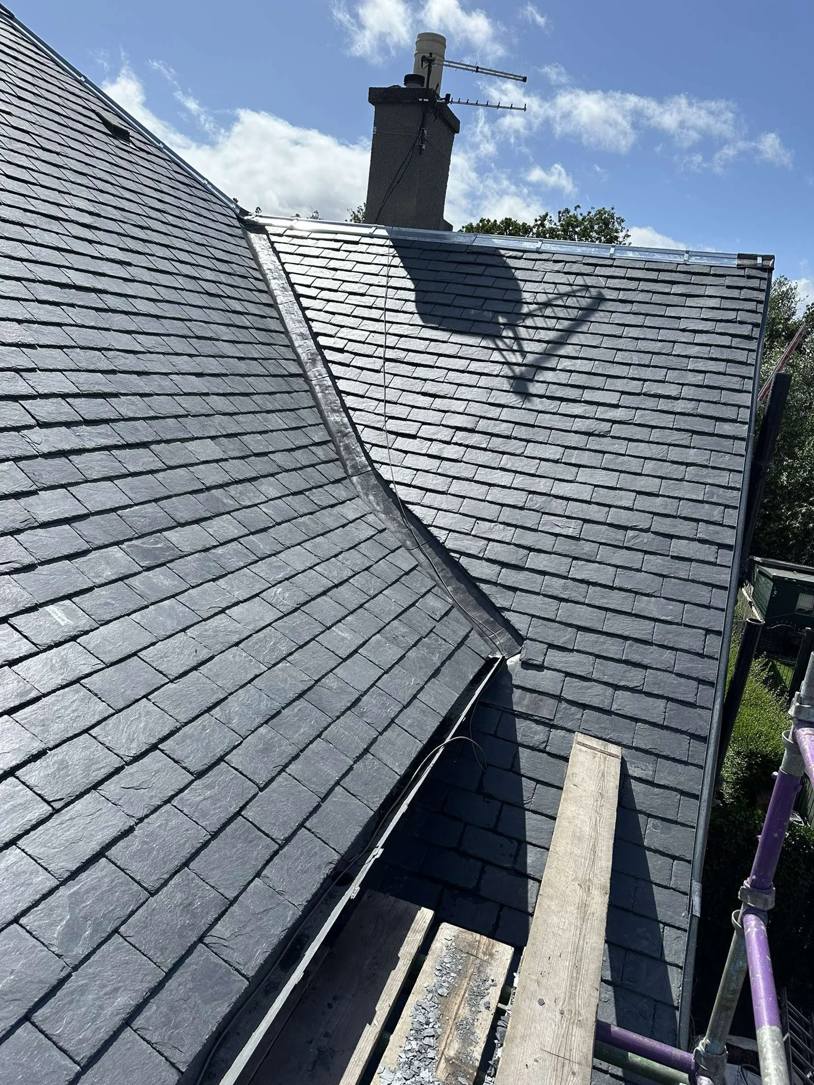 Photo of a rooftop with black slate shingles, a chimney with an antenna, and a wooden scaffold walkway.