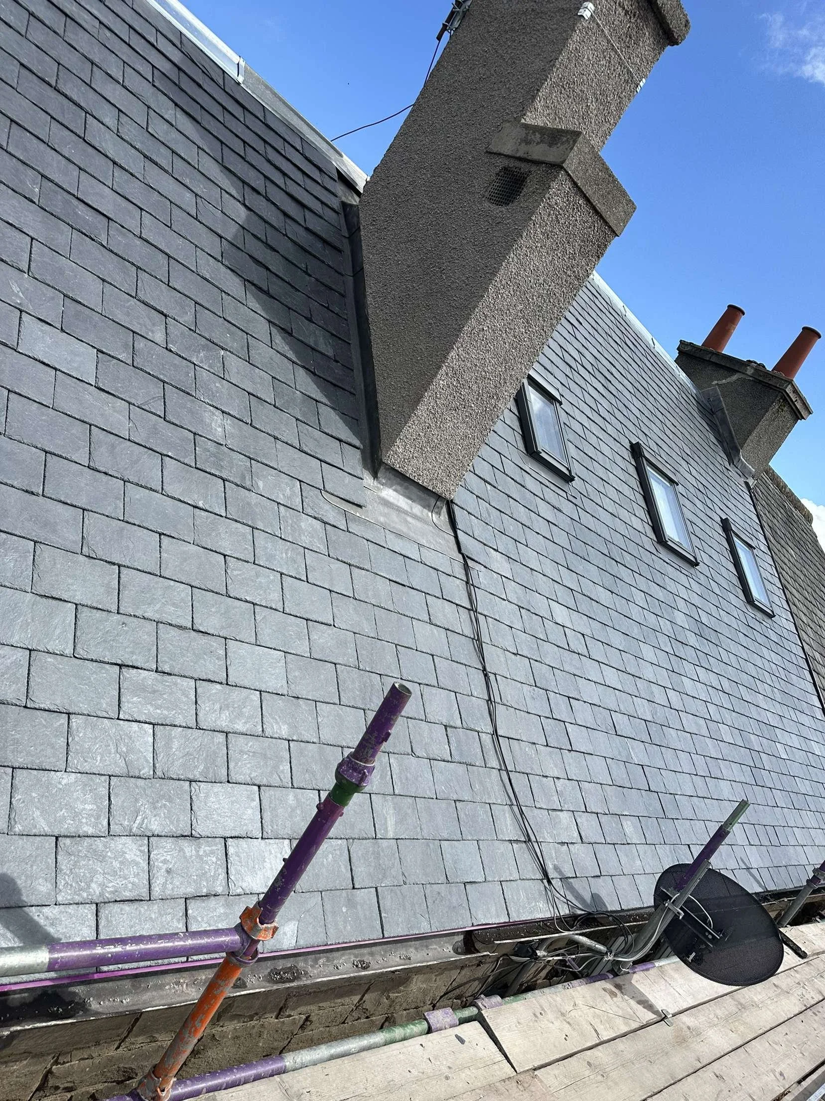 View of a building's slate roof with three skylights, a large chimney, and scaffolding on the edge, under a clear blue sky.
