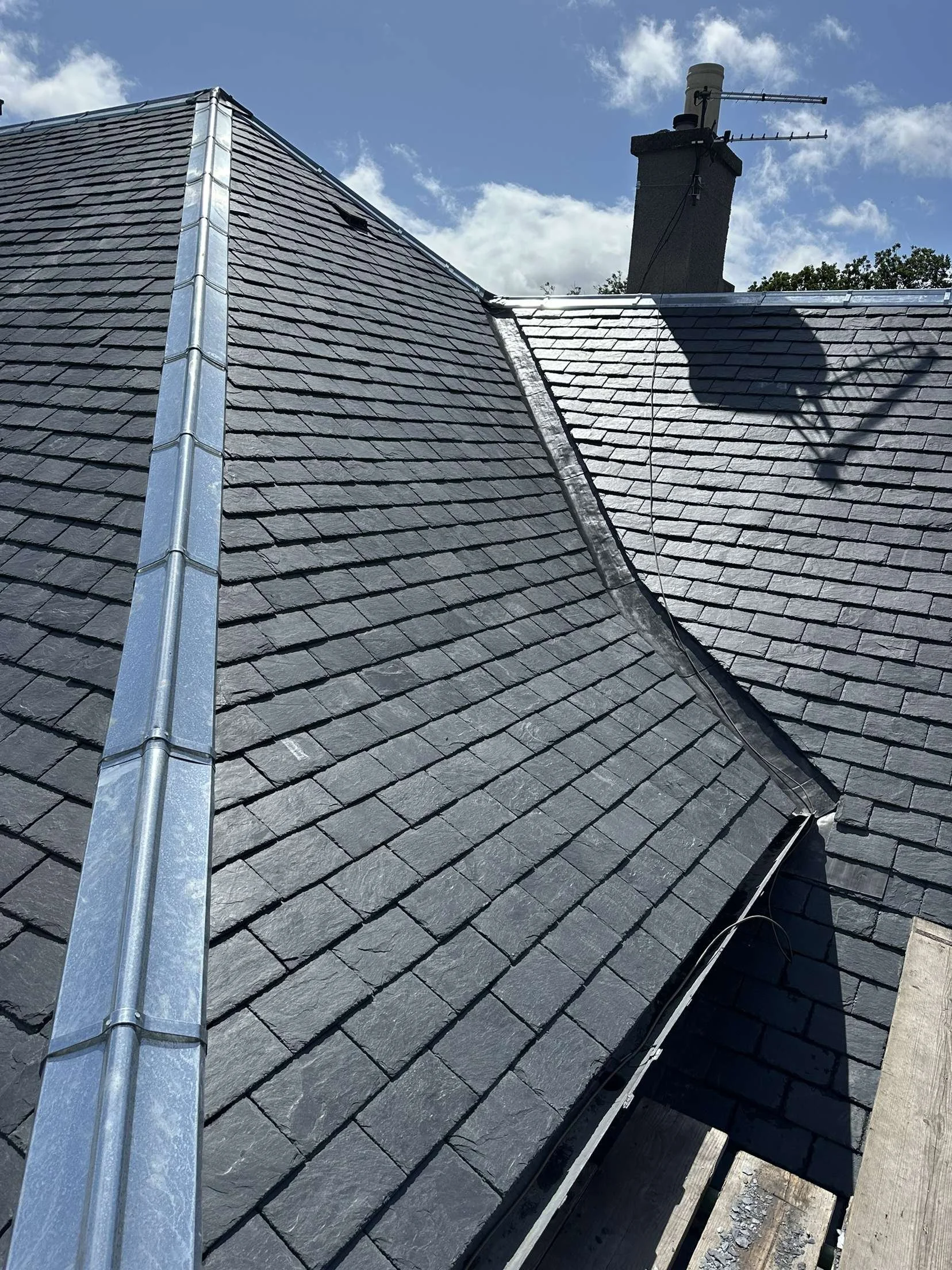 Close-up of a dark gray slate shingle roof with a metal flashing strip along the ridge, a chimney with an antenna, and a bright sky with some clouds.