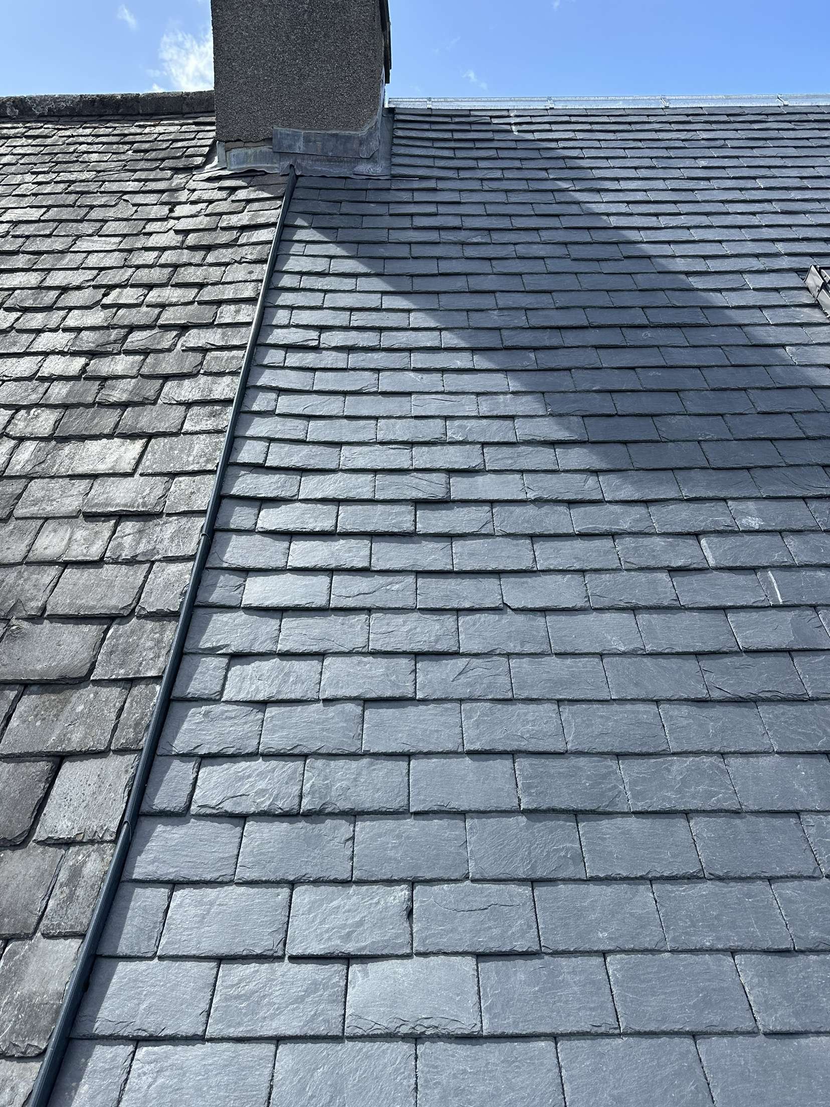 Close-up of a roof with black slate shingles, divided into two sections with different styles, under a blue sky with some clouds.