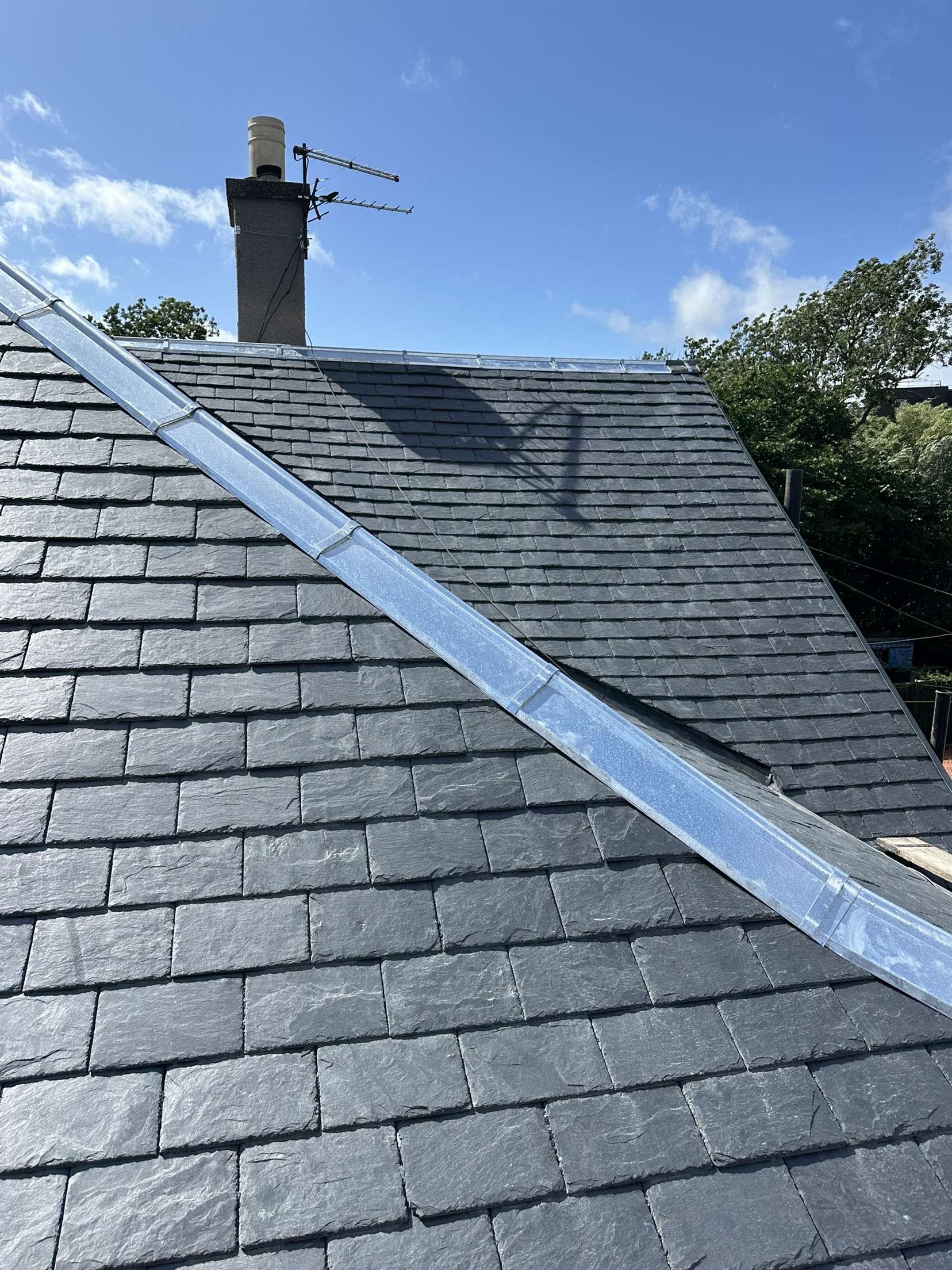 A close-up view of a house roof with gray slate shingles and a metal flashing strip running along the slope. In the background, there is a chimney with an antenna on top and a blue sky with some white clouds.
