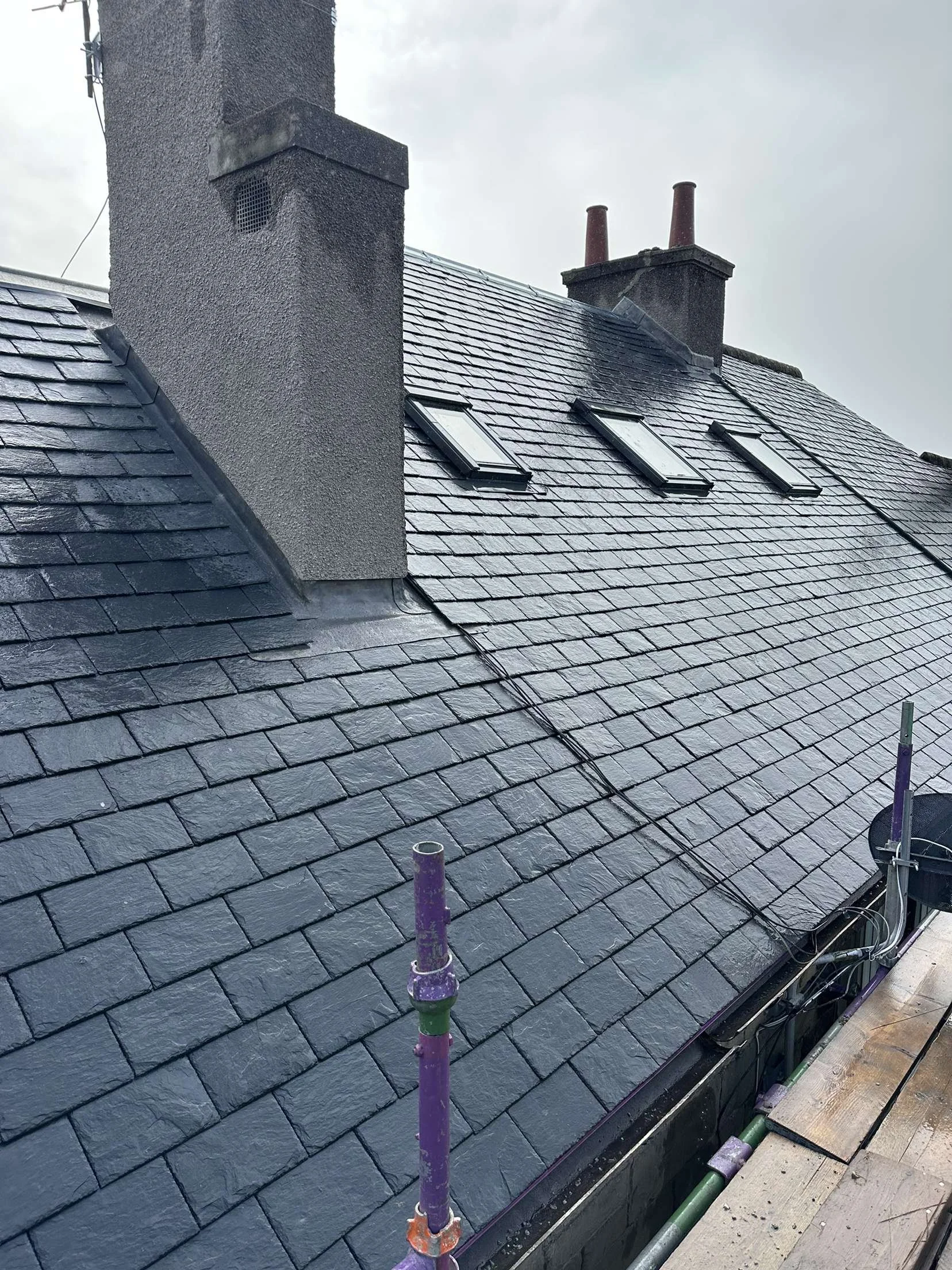 Roof with three skylights, chimneys, and wet slate shingles, construction scaffolding in the foreground.