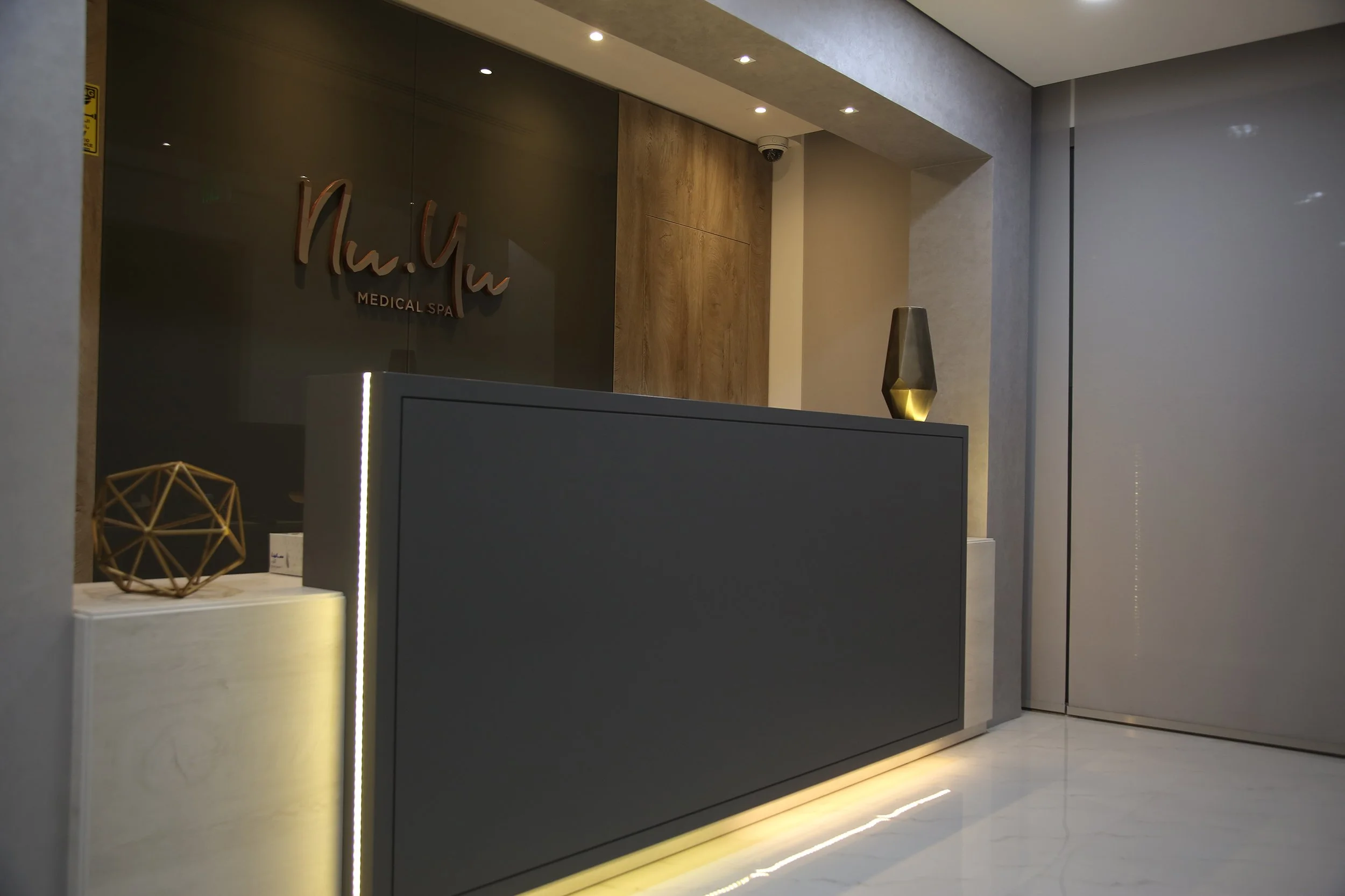 The sleek reception desk, illuminated by warm, inviting light, creates a sophisticated and welcoming atmosphere. The minimalist design with clean lines and subtle details reflects the clinic's focus on elegance and professionalism.