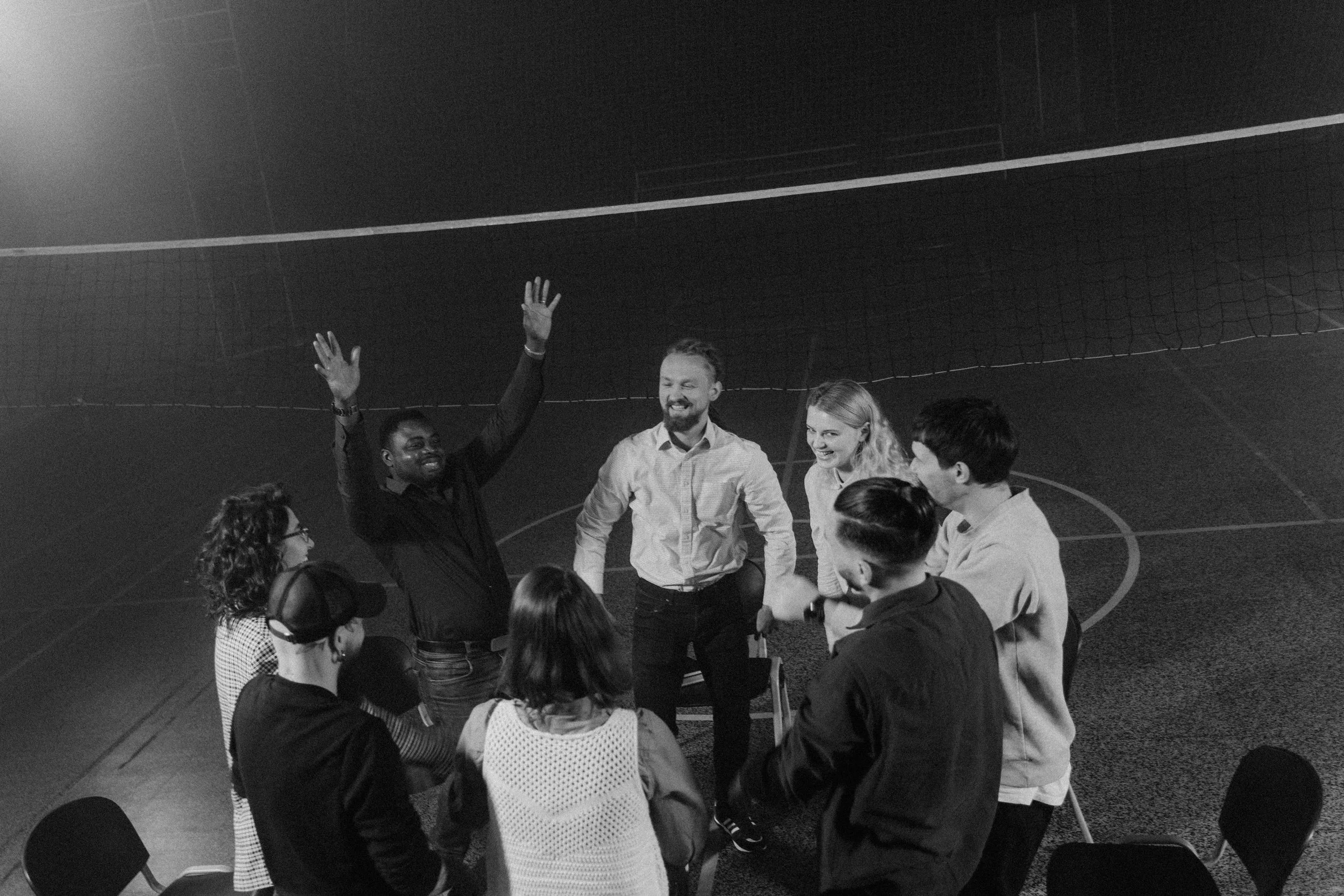 A group of diverse people standing in a circle and engaging in a discussion or activity in a gymnasium or sports hall. Some are smiling and raising their hands.