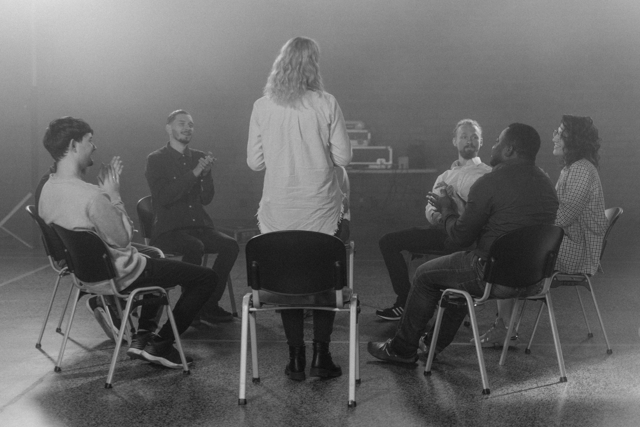 A group of people sitting in chairs in a circle, with one woman standing in the middle of the circle. They appear to be in a classroom or workshop setting, engaging in a discussion or activity.